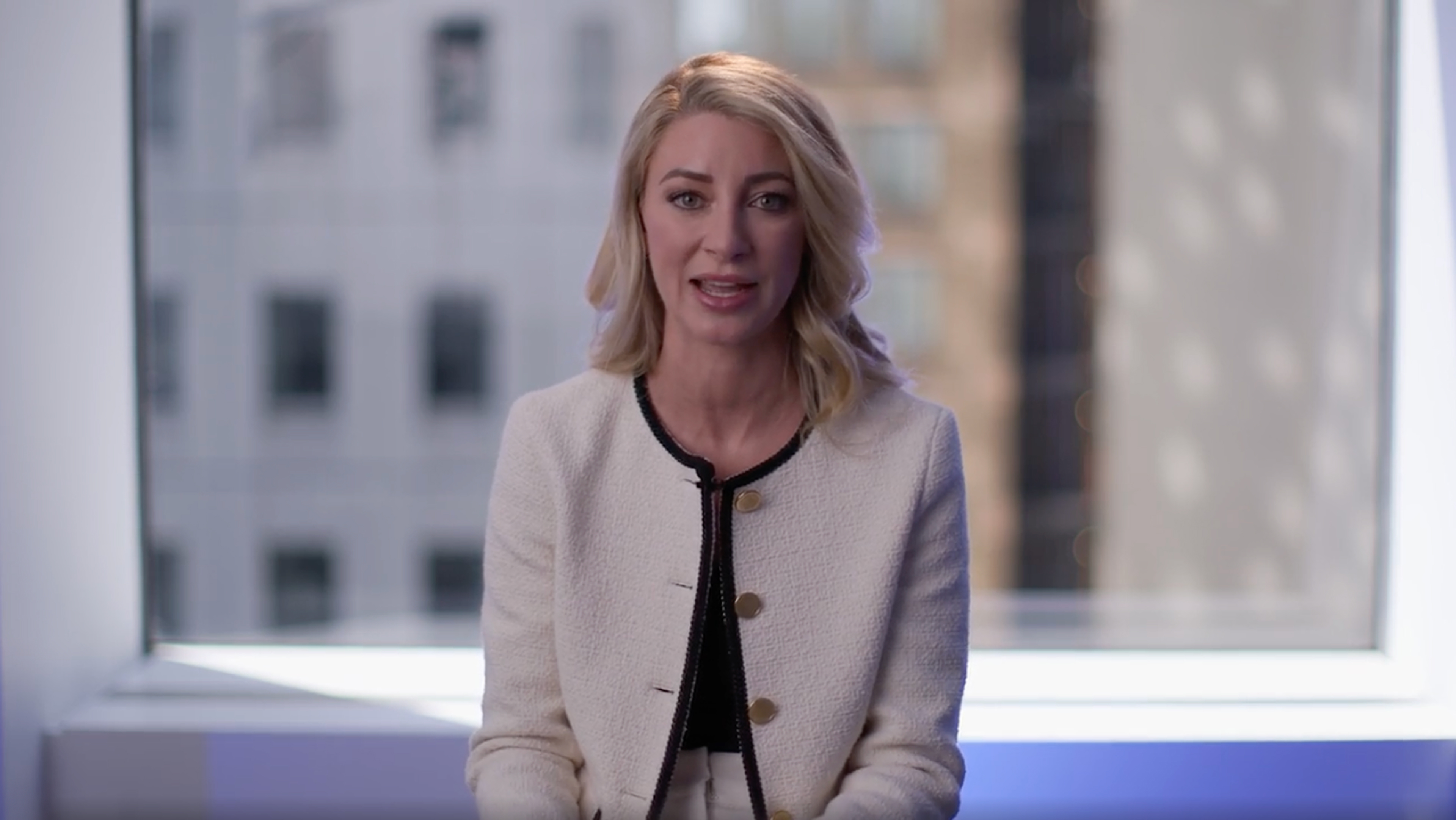 A person with shoulder-length blonde hair wearing a white blazer with gold buttons and black trim, seated indoors near a window with blurred city buildings outside.