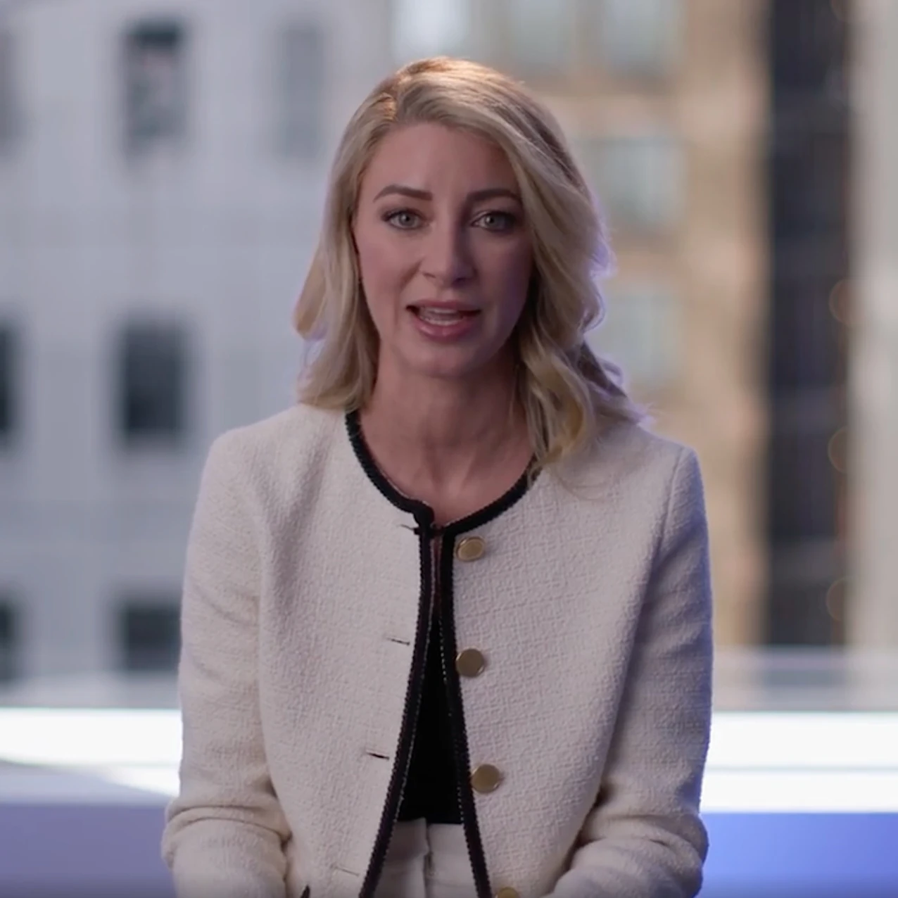 A person with shoulder-length blonde hair wearing a white blazer with gold buttons and black trim, seated indoors near a window with blurred city buildings outside.