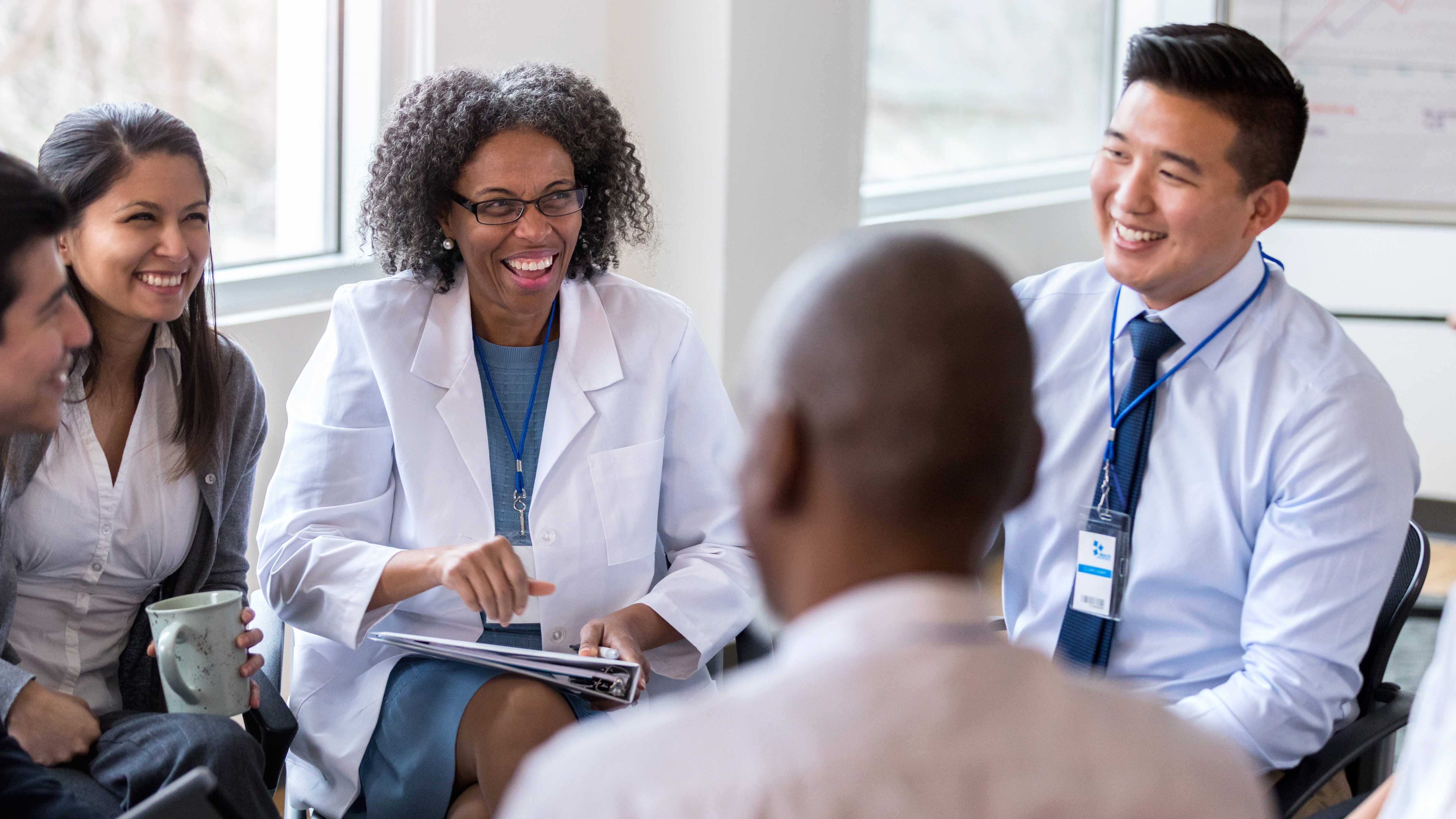 A diverse group of five professionals engaged in a lively discussion in a bright office, with one woman holding a clipboard and smiling.