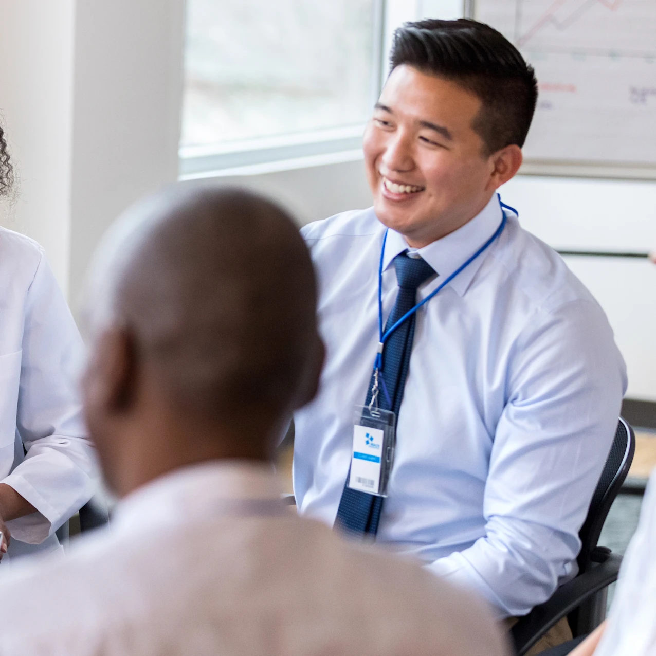 A diverse group of five professionals engaged in a lively discussion in a bright office, with one woman holding a clipboard and smiling.