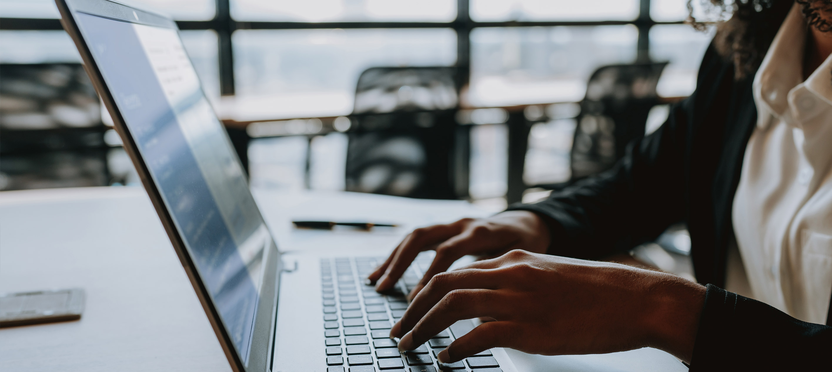 Close-up of hands typing on a laptop keyboard at a desk, with a phone, pen and a person seated nearby.