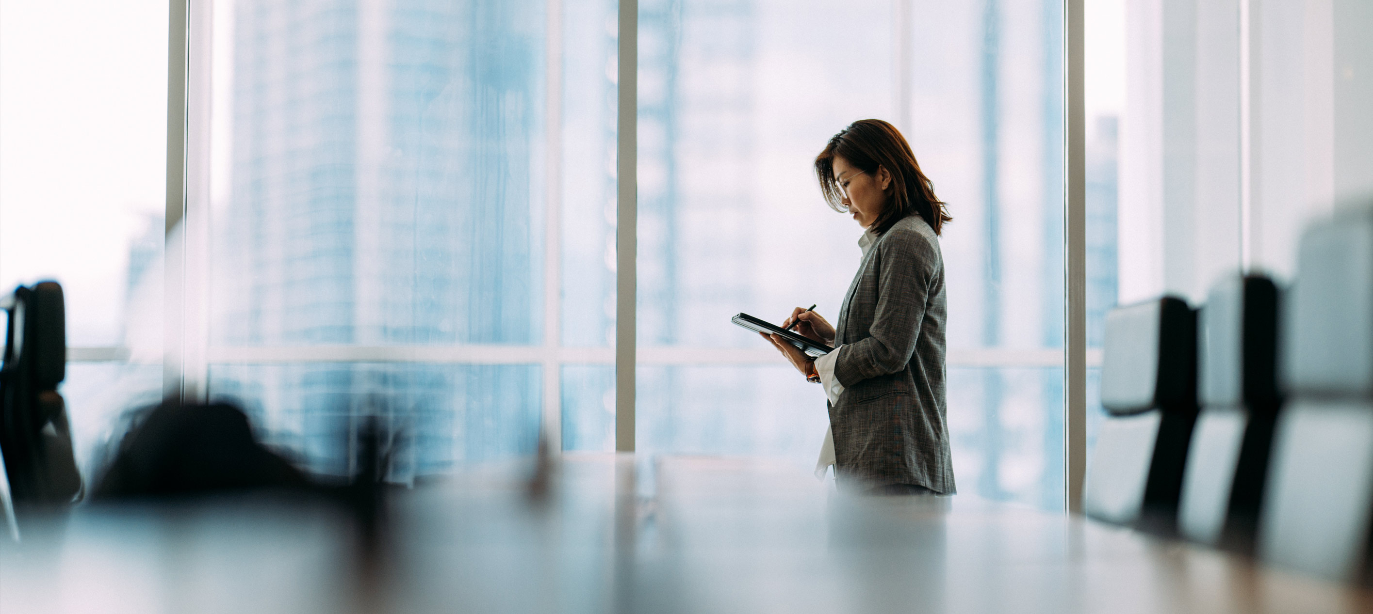 Woman in a plaid blazer stands writing on a tablet in a glass-walled office with city skyscrapers visible.