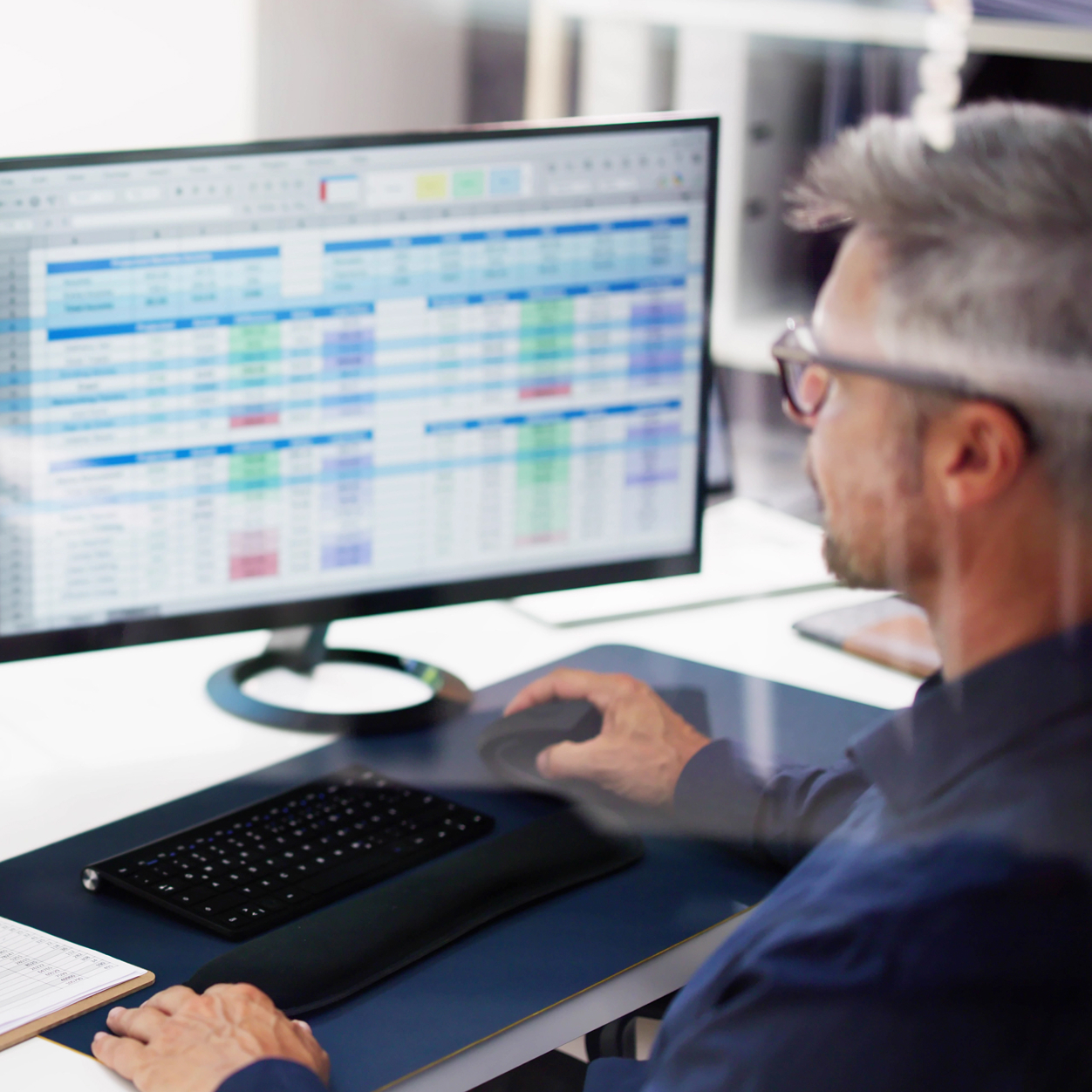 Man working at a desk using a computer mouse with a large monitor displaying spreadsheets with rows and columns of data in various colors.