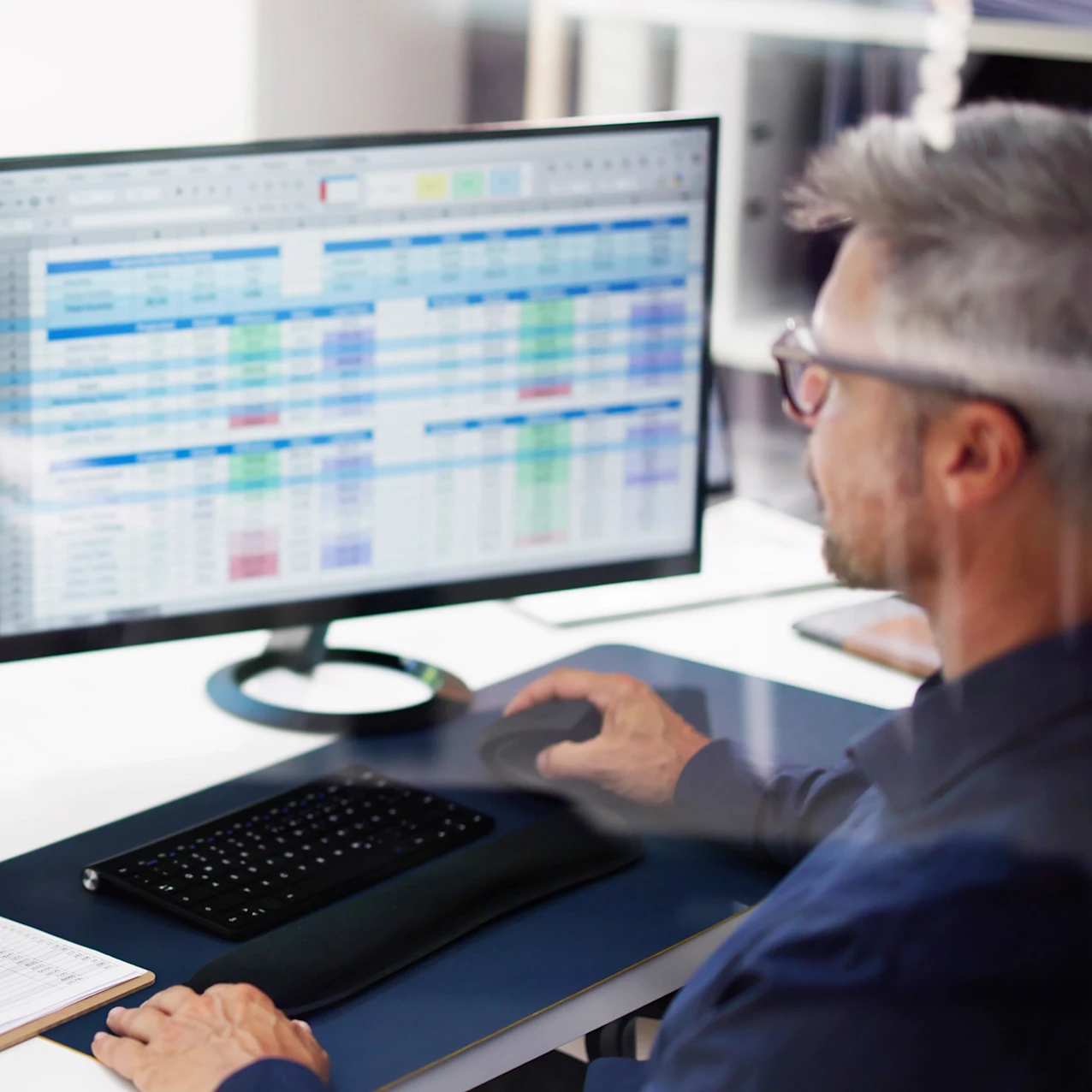 Man working at a desk using a computer mouse with a large monitor displaying spreadsheets with rows and columns of data in various colors.