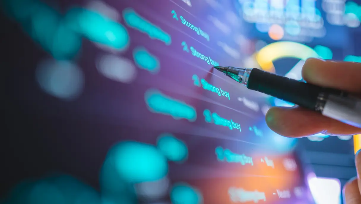 Close-up of a hand holding a pen while analyzing digital stock market data, trading signals and financial charts displayed on a screen.