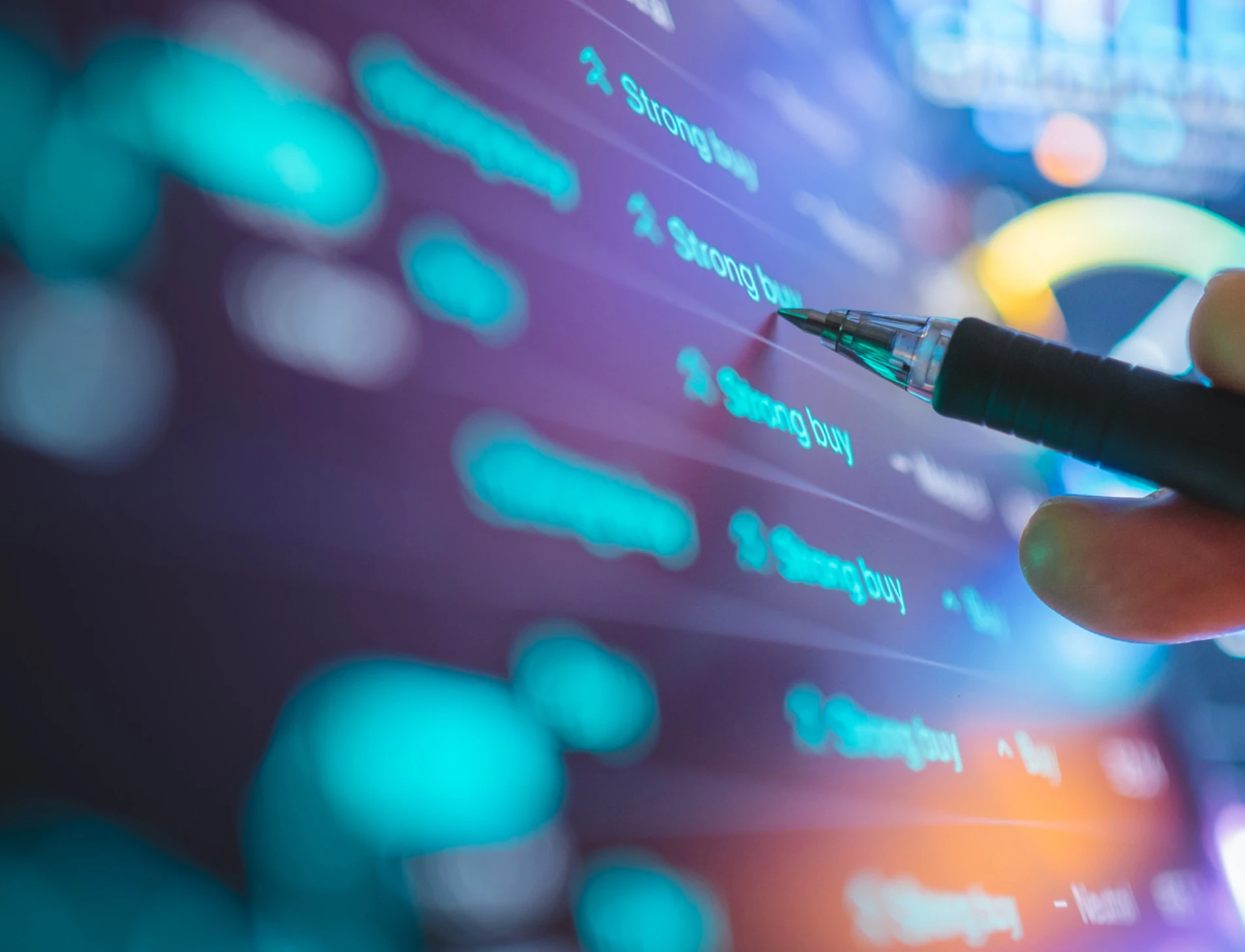 Close-up of a hand holding a pen while analyzing digital stock market data, trading signals and financial charts displayed on a screen.