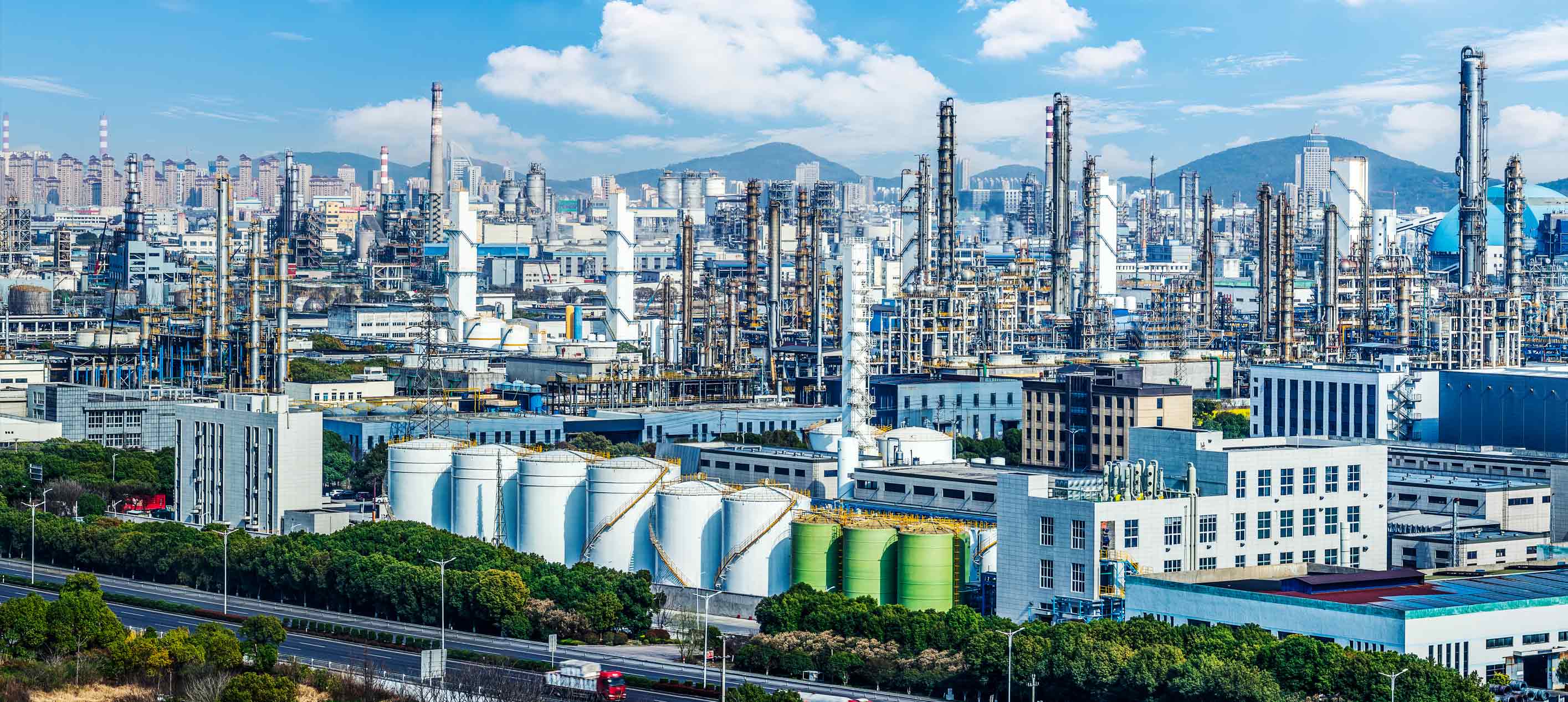 Sprawling industrial complex with refinery towers, storage tanks and factory buildings against a city skyline.