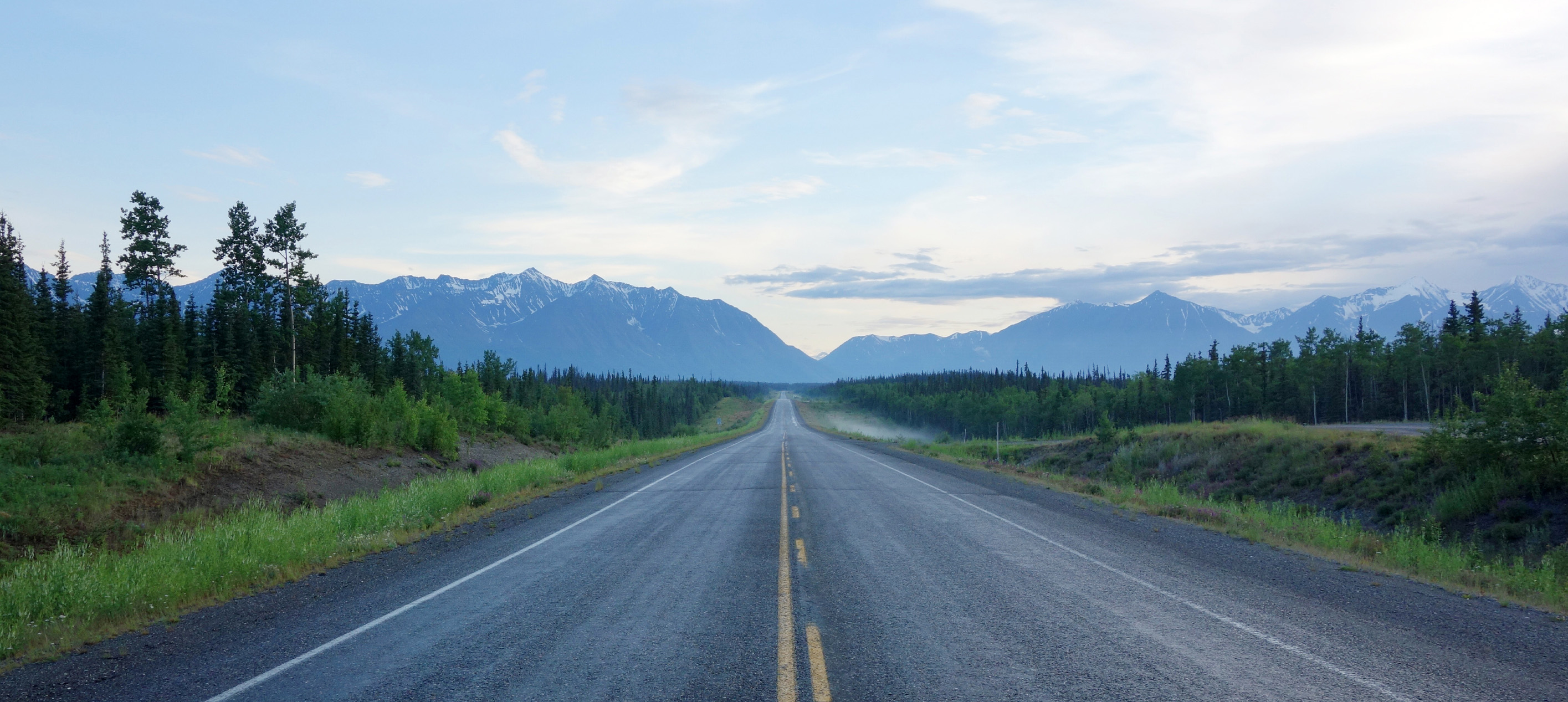 Long empty highway stretching through a forest toward distant mountain ranges beneath a partly cloudy sky.