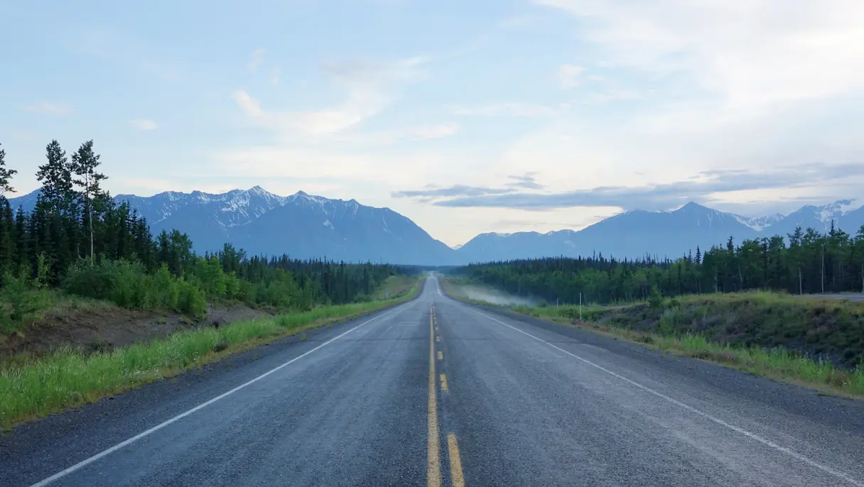 Long empty highway stretching through a forest toward distant mountain ranges beneath a partly cloudy sky.