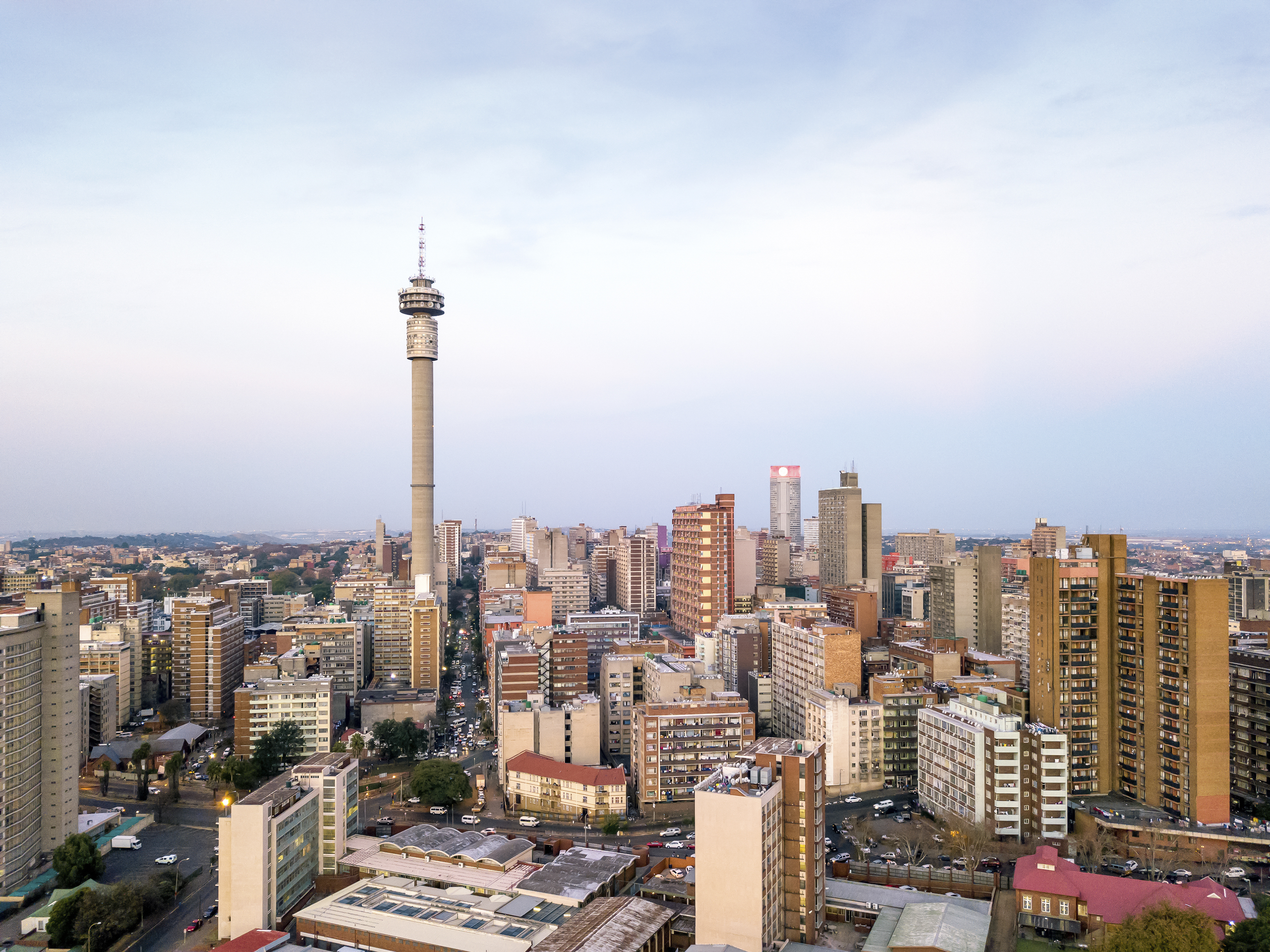 A cityscape featuring numerous mid-rise buildings with a prominent tall telecommunications tower in the center under a clear sky at dusk.