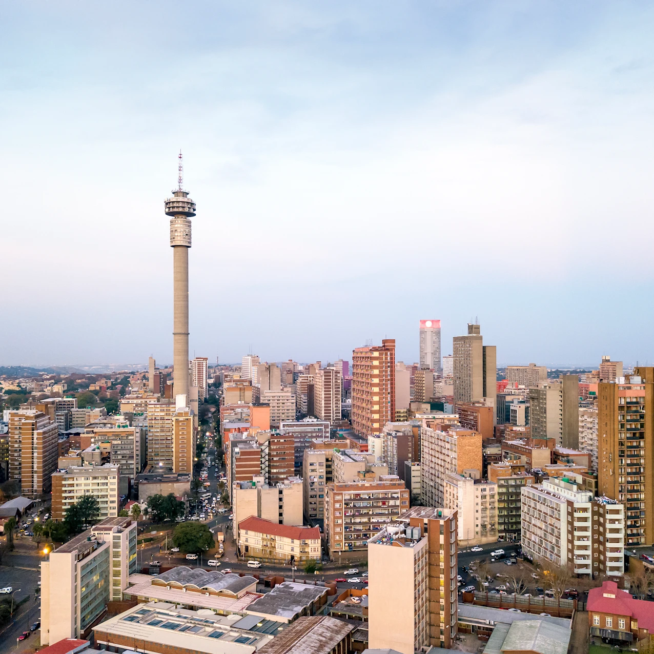 A cityscape featuring numerous mid-rise buildings with a prominent tall telecommunications tower in the center under a clear sky at dusk.