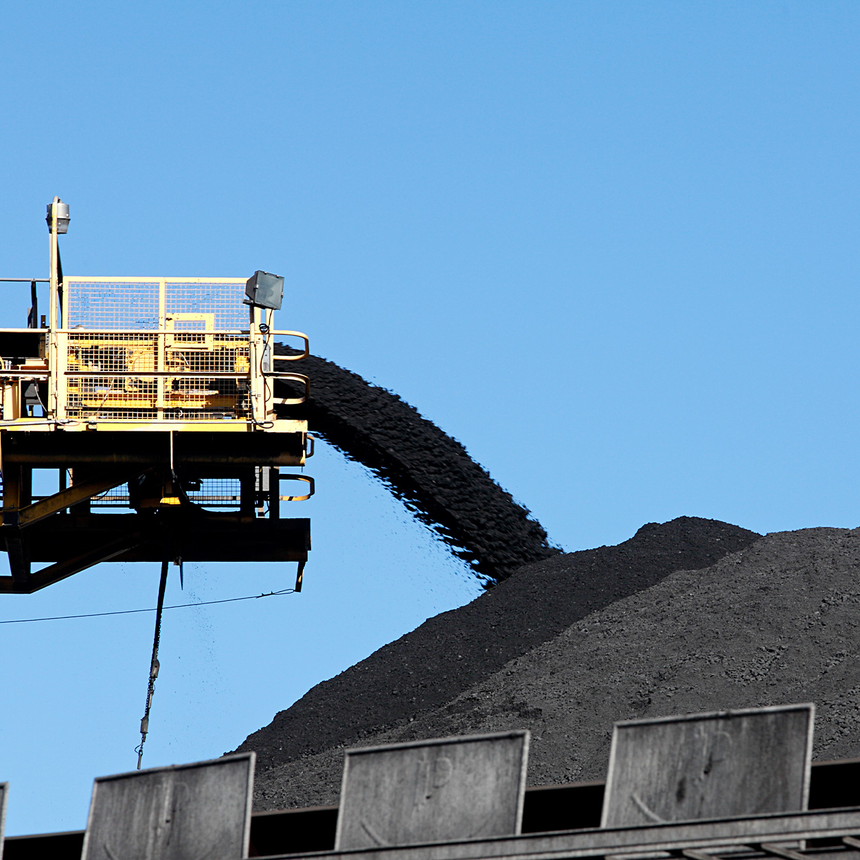 Black coal is poured from a conveyor belt onto a large pile under a clear blue sky at an industrial site.