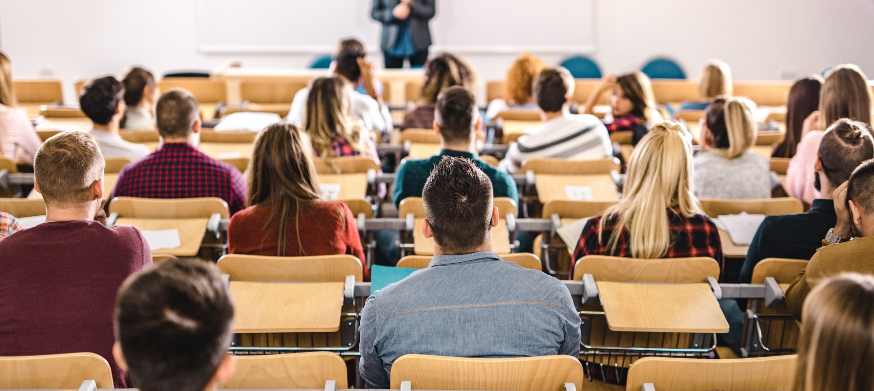 university or college students seated at wooden desks in a tiered lecture hall, facing a professor standing at the front near a whiteboard.
