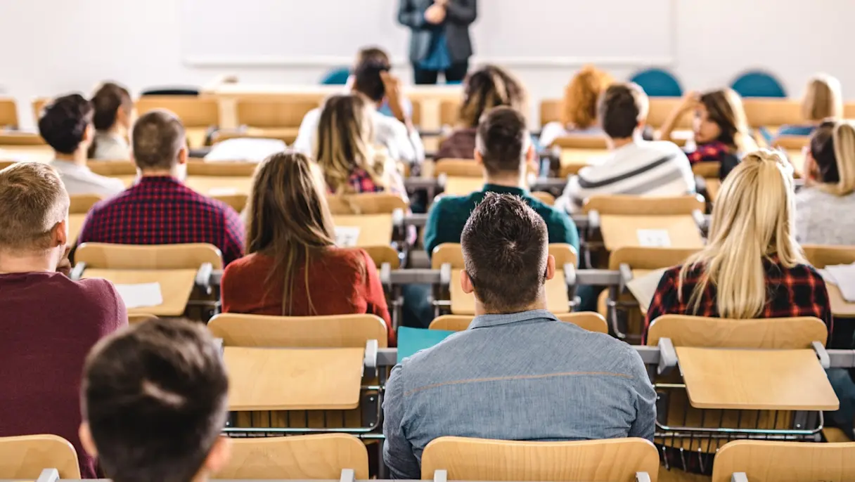 university or college students seated at wooden desks in a tiered lecture hall, facing a professor standing at the front near a whiteboard.
