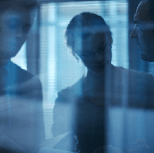 Two blurred people behind a glass wall with reflections, in a dimly lit office space with vertical blinds.