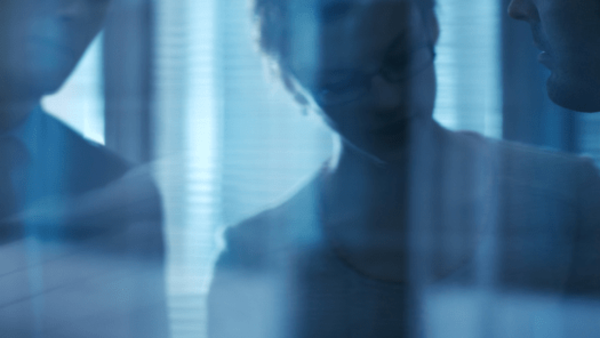 Two blurred people behind a glass wall with reflections, in a dimly lit office space with vertical blinds.