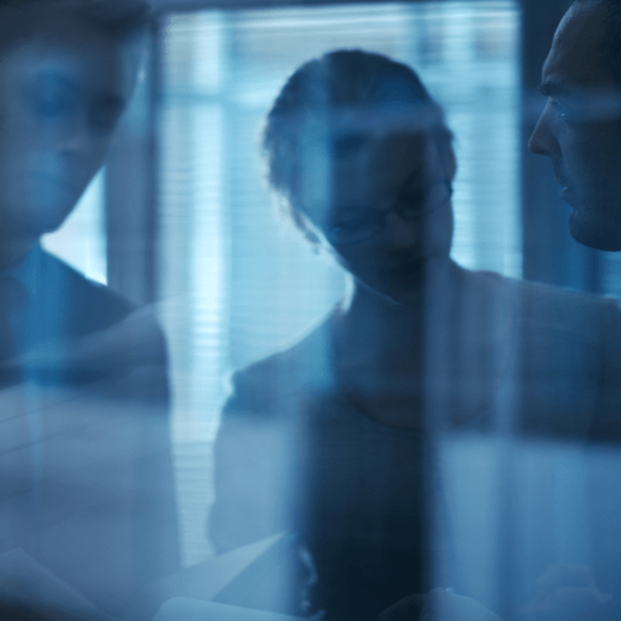 Two blurred people behind a glass wall with reflections, in a dimly lit office space with vertical blinds.