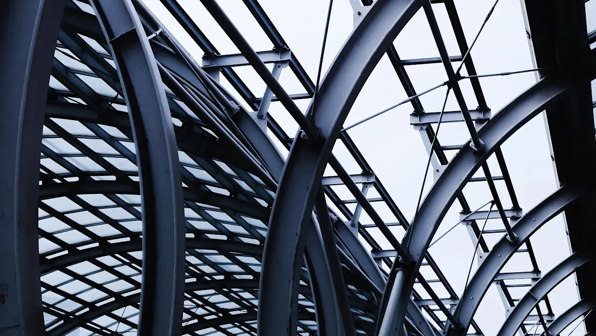 Upward view of a modern glass roof supported by curved steel beams and an intricate geometric framework.