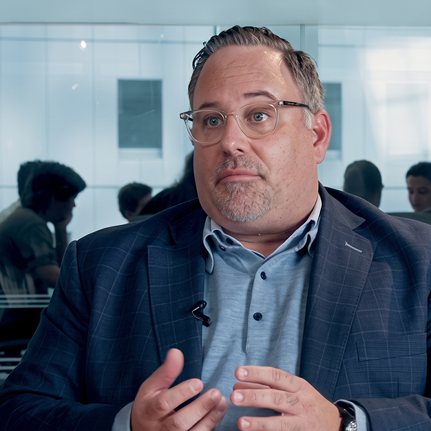 Middle-aged man with glasses and short hair wearing a blue checkered blazer and light blue shirt speaks with hand gestures in a modern office with people blurred in the background.
