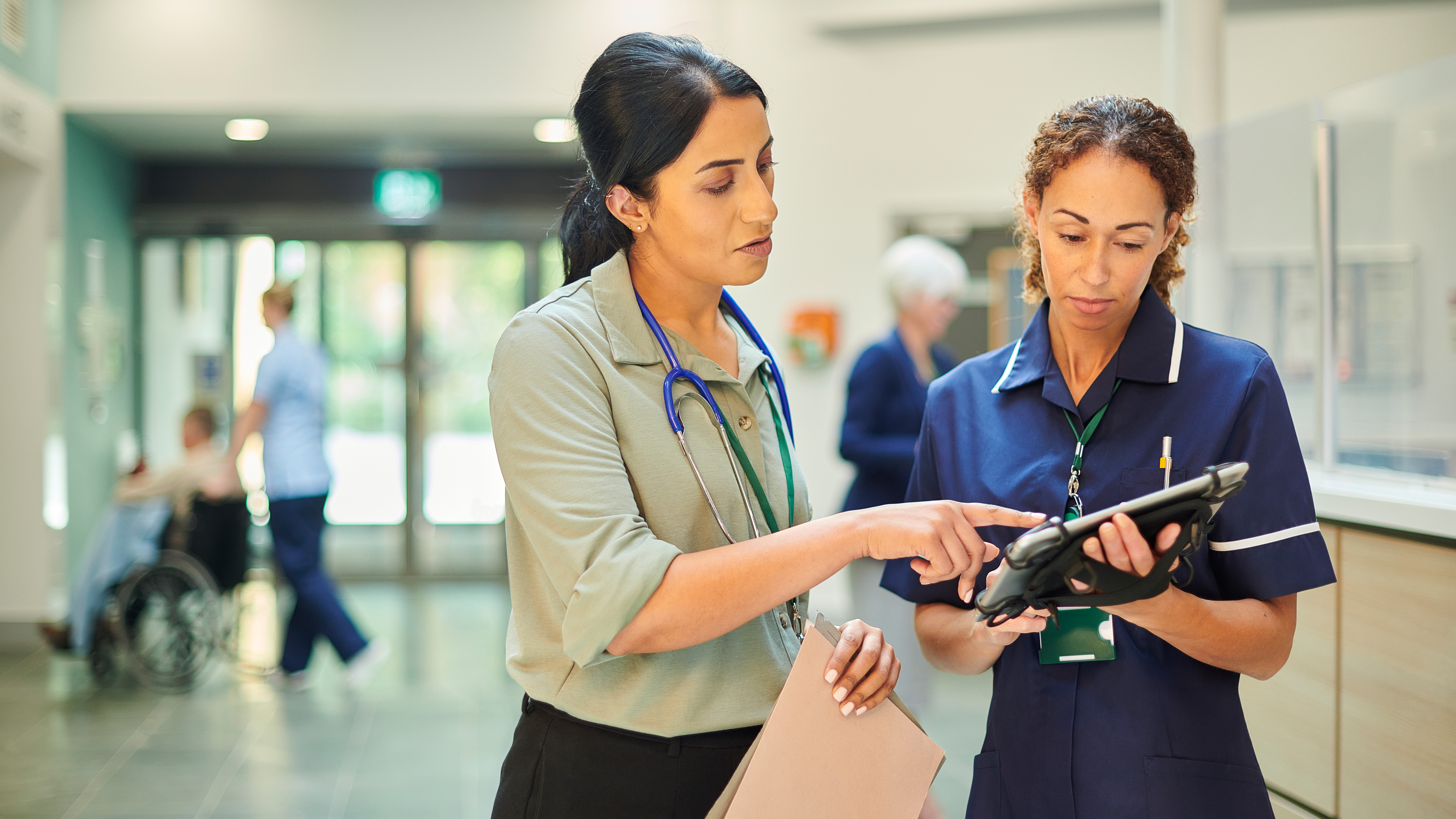 Two healthcare professionals in a hospital corridor, one pointing at a tablet held by the other, with a wheelchair and staff visible in the background.