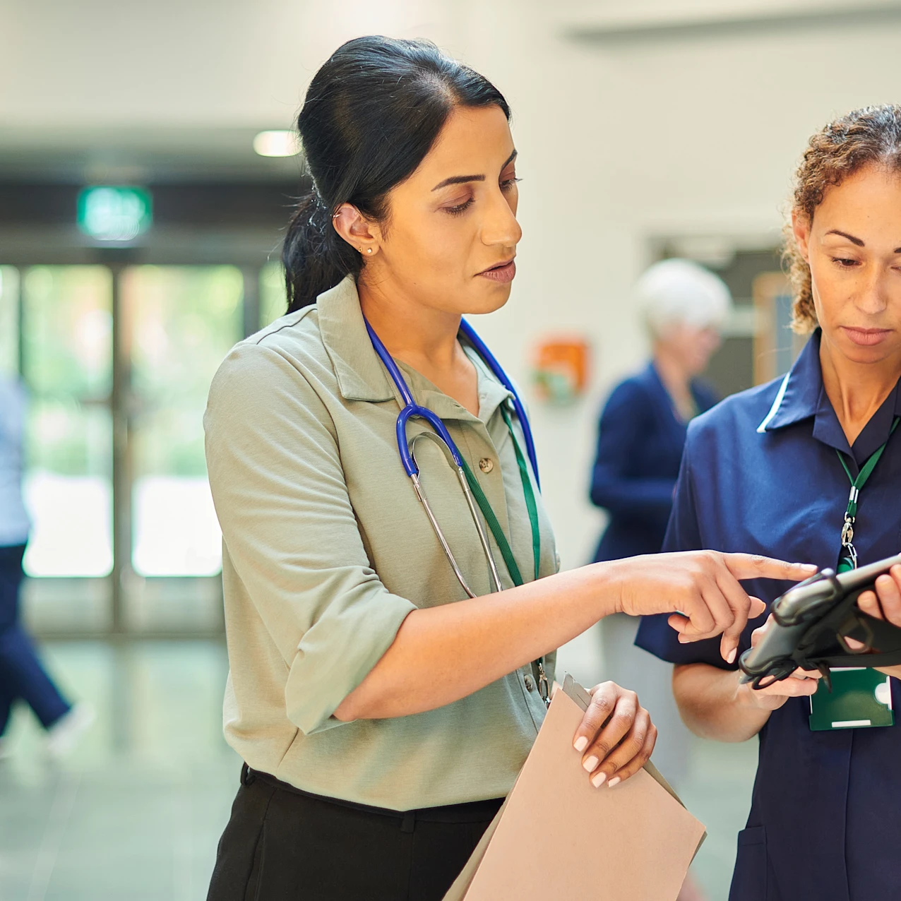 Two healthcare professionals in a hospital corridor, one pointing at a tablet held by the other, with a wheelchair and staff visible in the background.