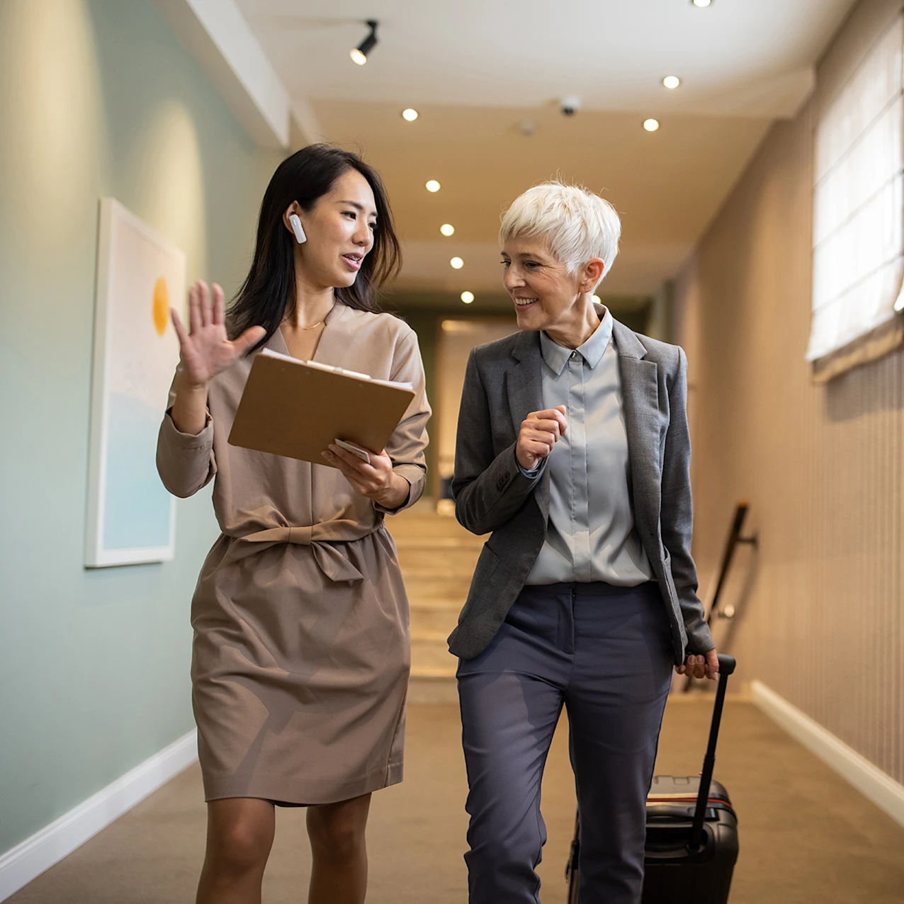 Two businesswomen walking down a corridor, one holding a tablet and gesturing while the other pulls a suitcase and listens.