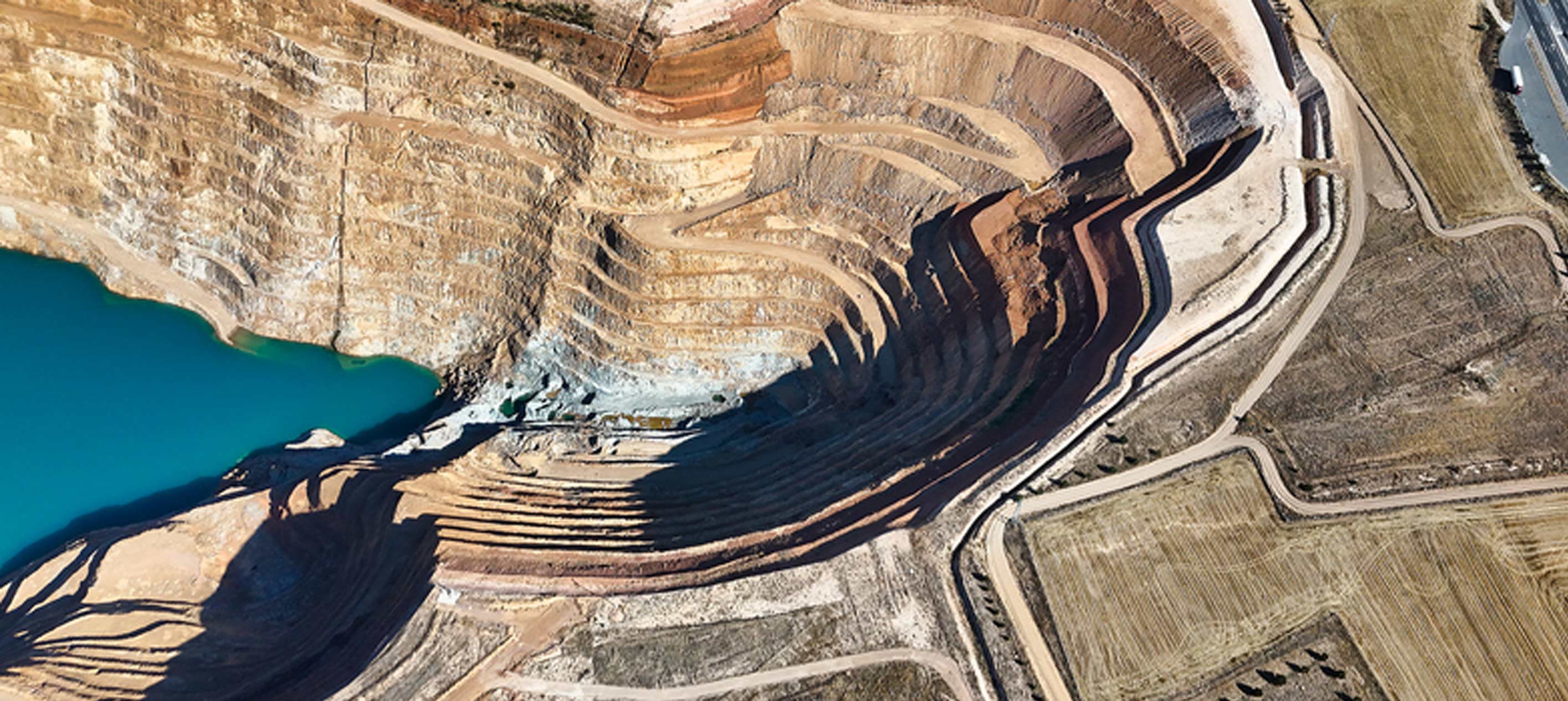 Aerial view of an open-pit mine with terraced excavation walls, winding access roads and a turquoise water reservoir.