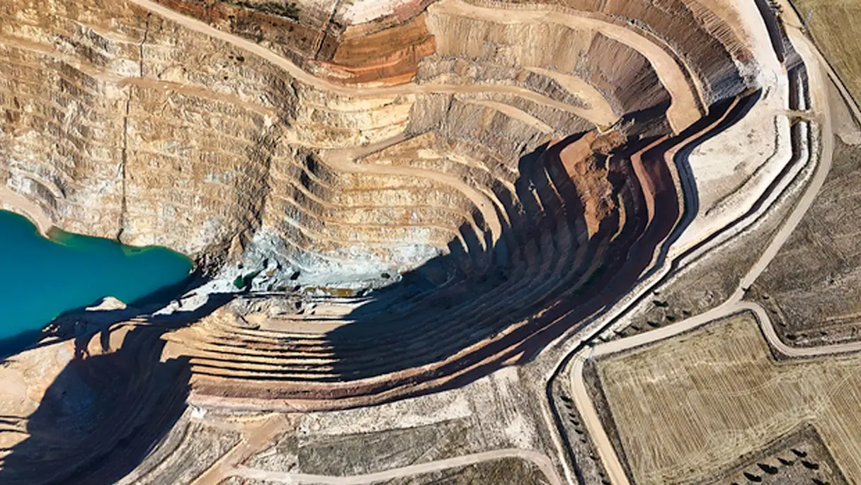 Aerial view of an open-pit mine with terraced excavation walls, winding access roads and a turquoise water reservoir.