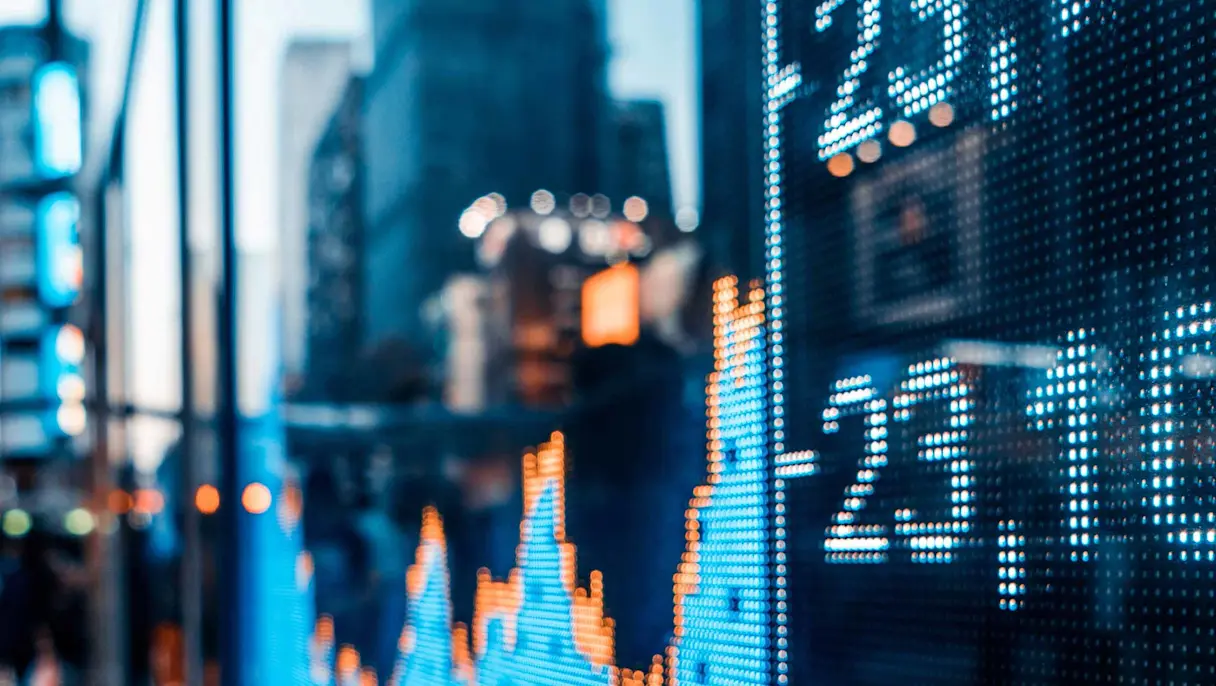 A digital display showing financial market data and graphs with blurred city buildings and lights visible in the background.