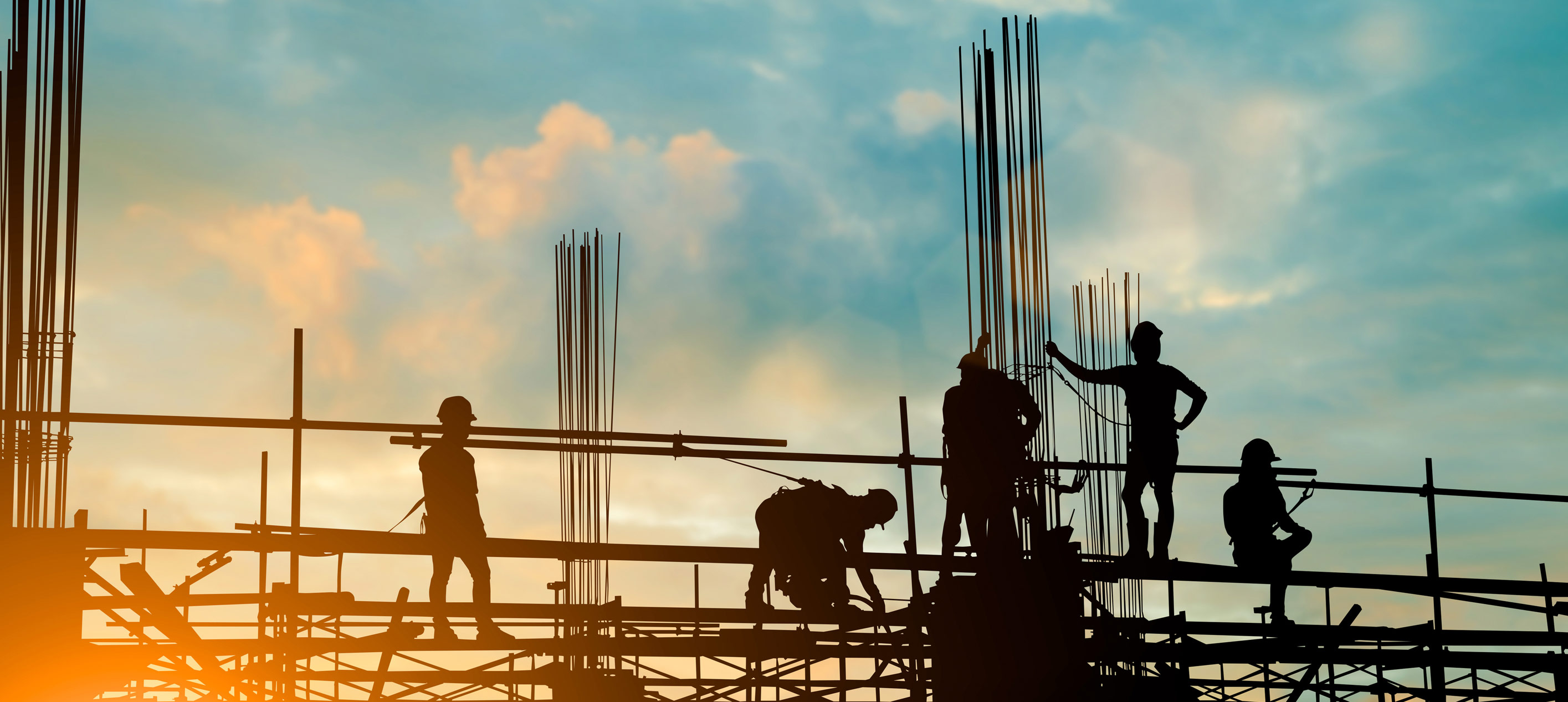 Silhouettes of construction workers on scaffolding with steel rods against a blue sky and warm orange sunset glow.