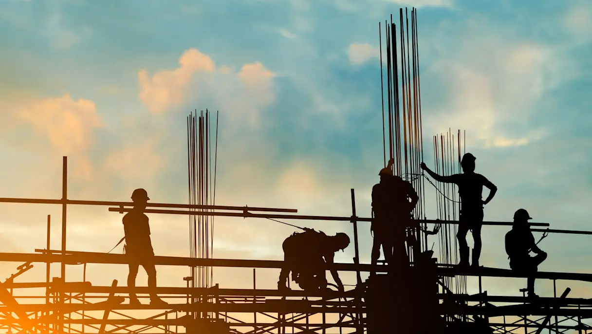 Silhouettes of construction workers on scaffolding with steel rods against a blue sky and warm orange sunset glow.