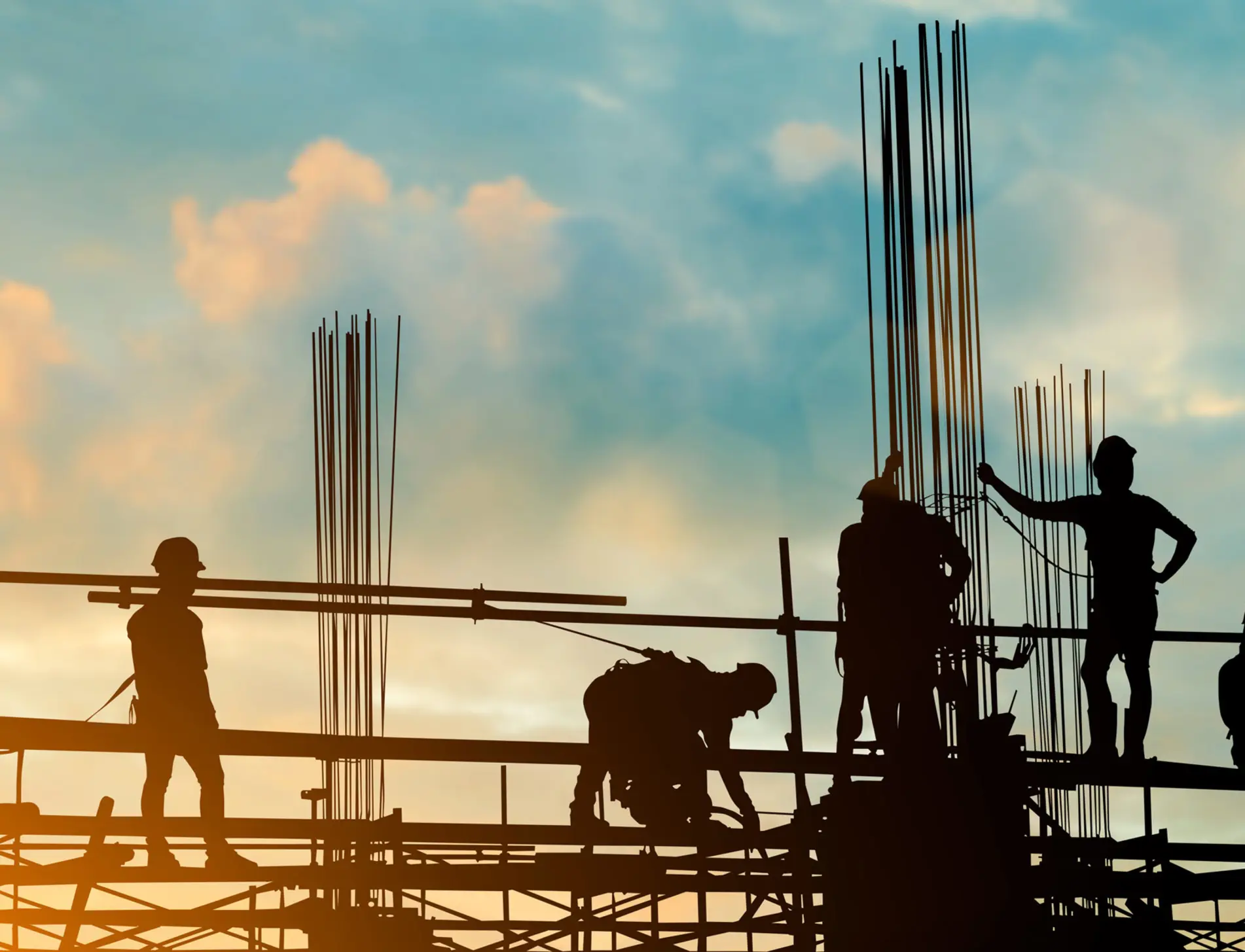 Silhouettes of construction workers on scaffolding with steel rods against a blue sky and warm orange sunset glow.
