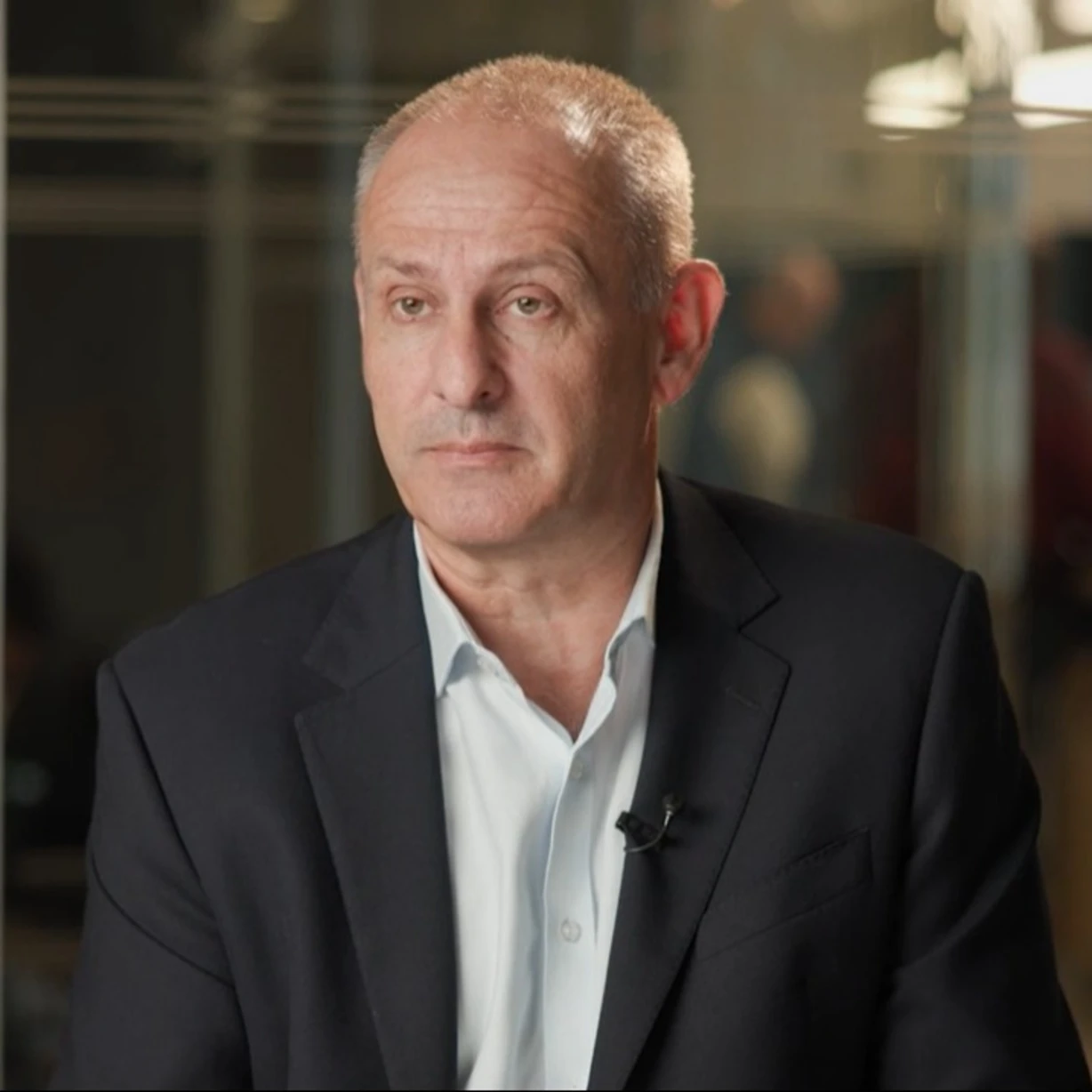 A person in a black suit and white shirt sitting indoors with a blurred background of people and lights.