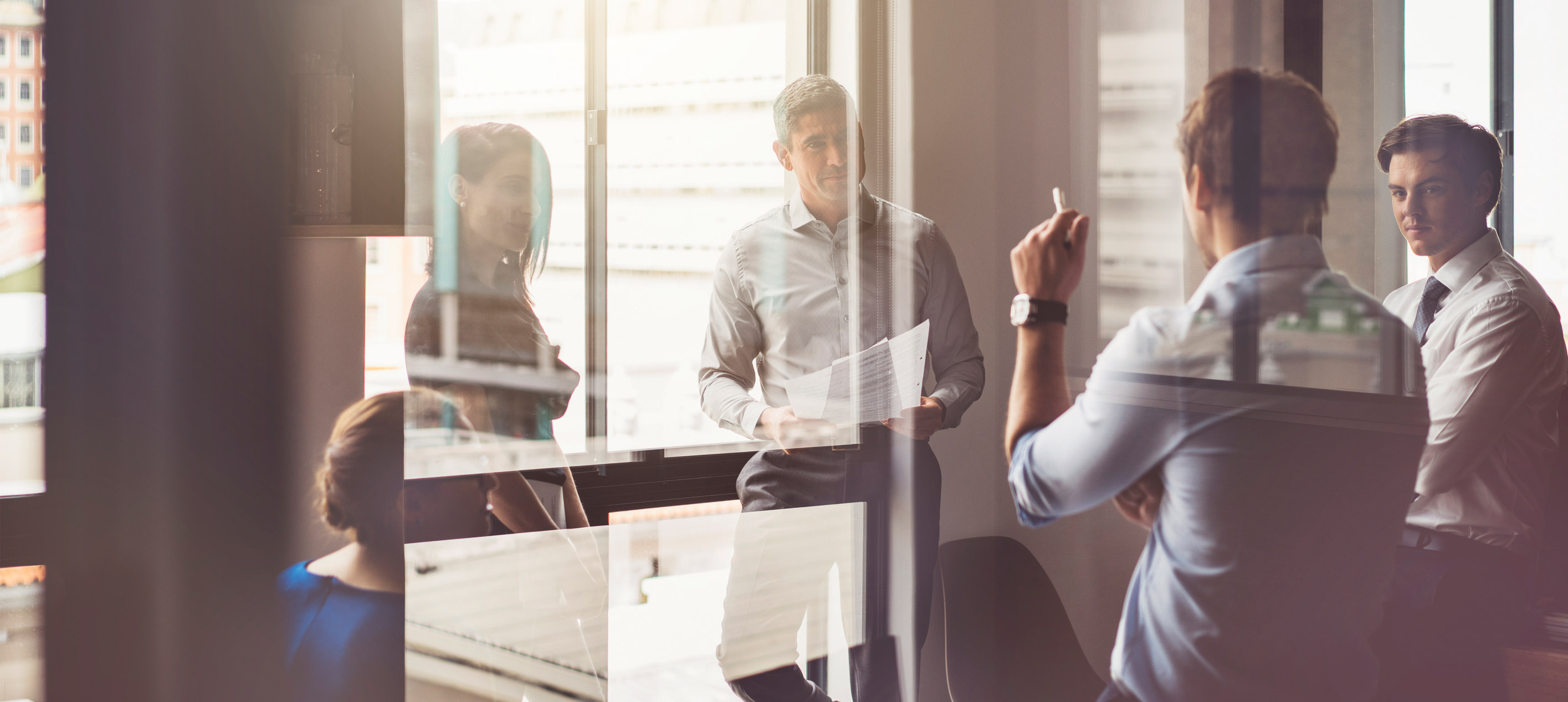A man holding papers speaks to colleagues in a glass-walled office, with one person raising a hand to ask something.