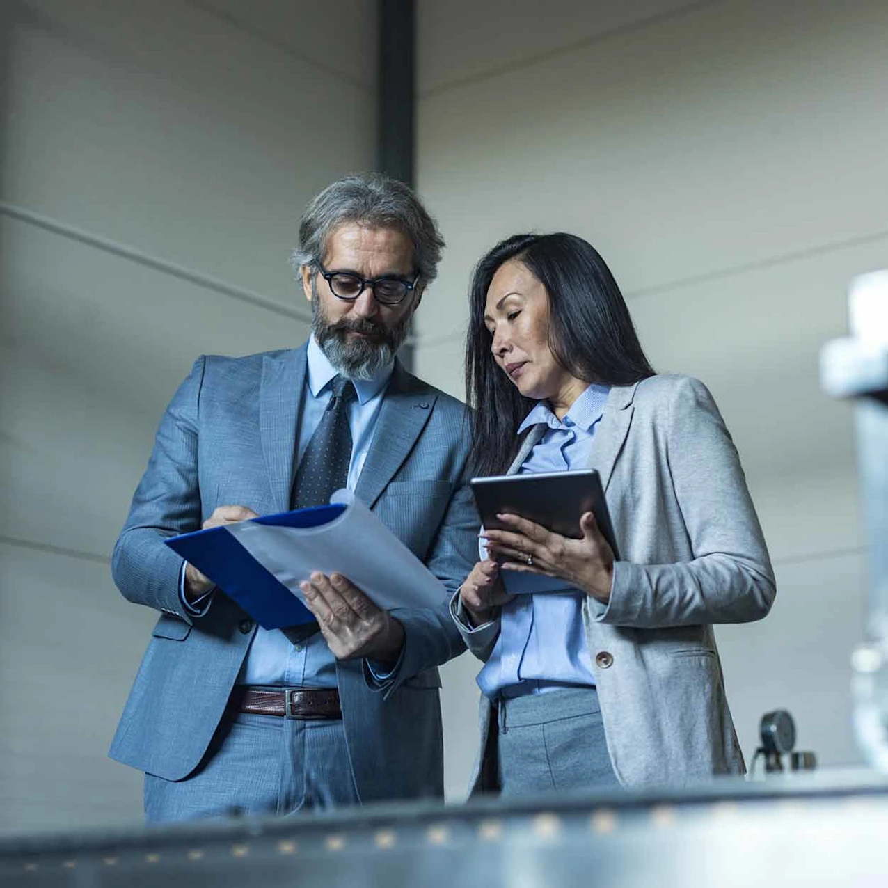 Two business professionals in gray suits review documents and use a tablet in an industrial setting with equipment in the foreground.
