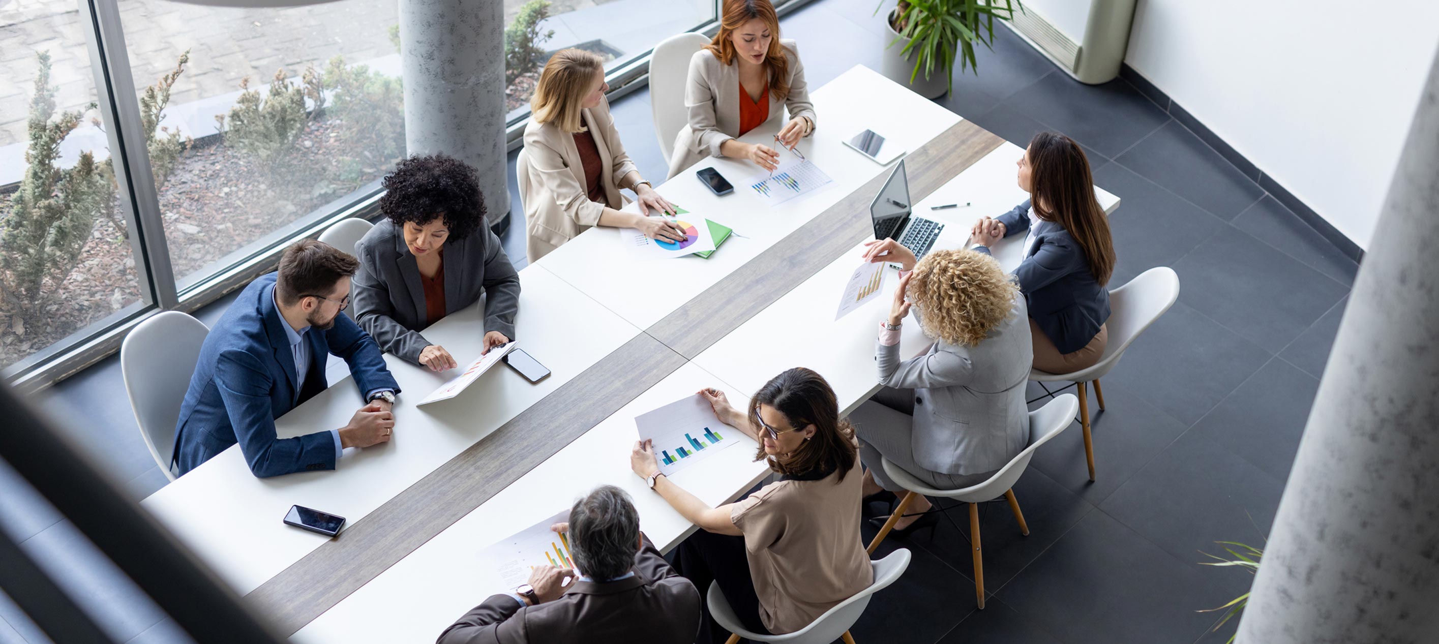 Overhead view of eight professionals seated around a long conference table reviewing printed charts and documents.