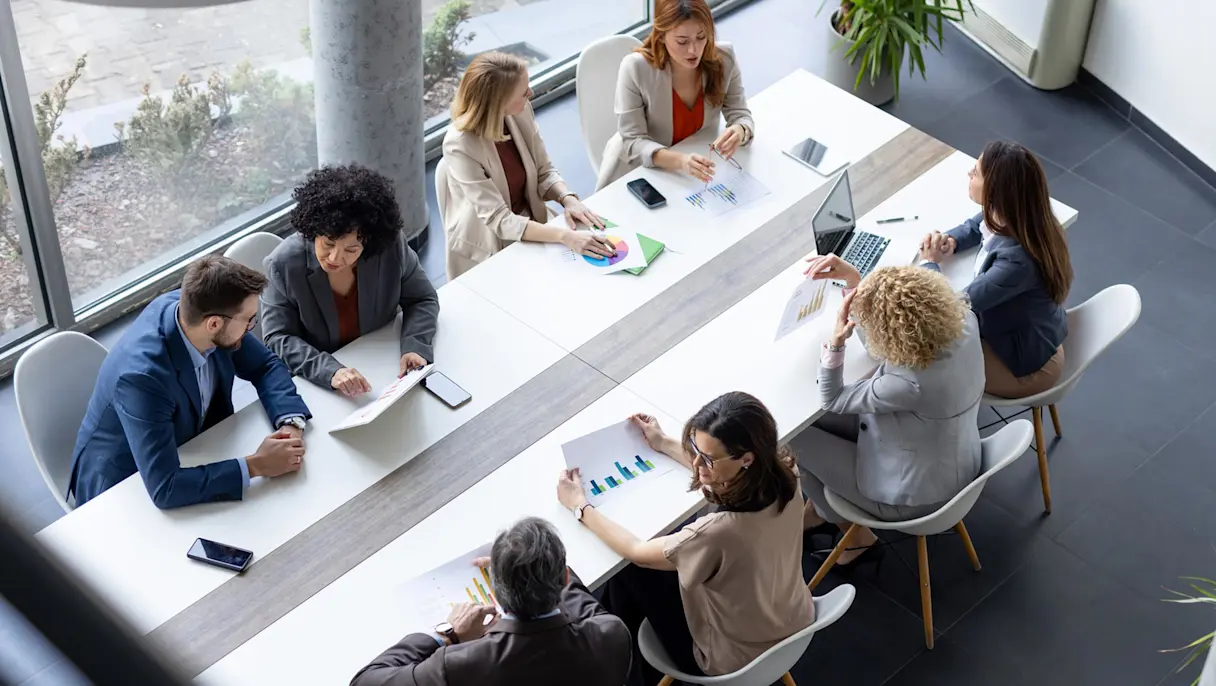 Overhead view of eight professionals seated around a long conference table reviewing printed charts and documents.