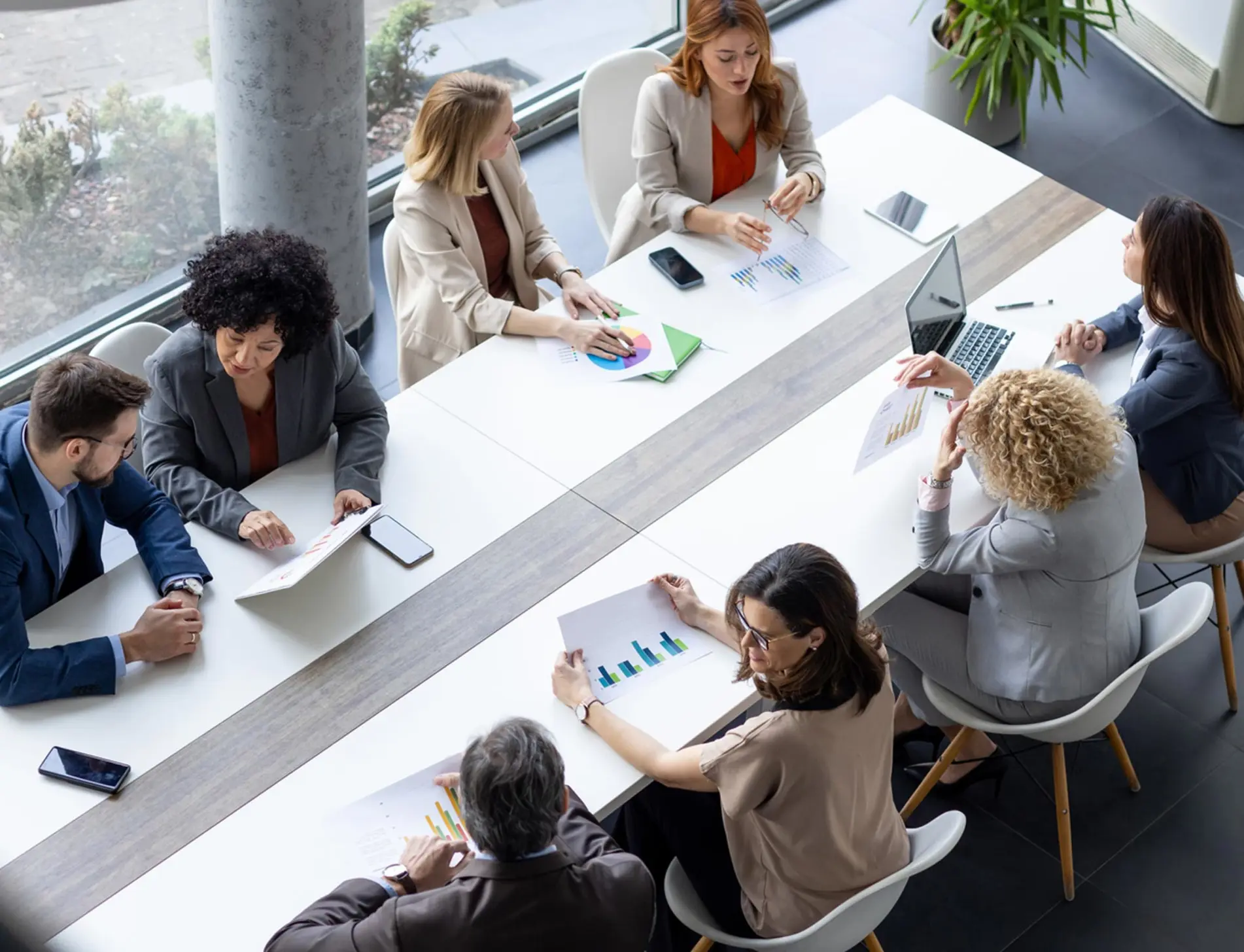 Overhead view of eight professionals seated around a long conference table reviewing printed charts and documents.