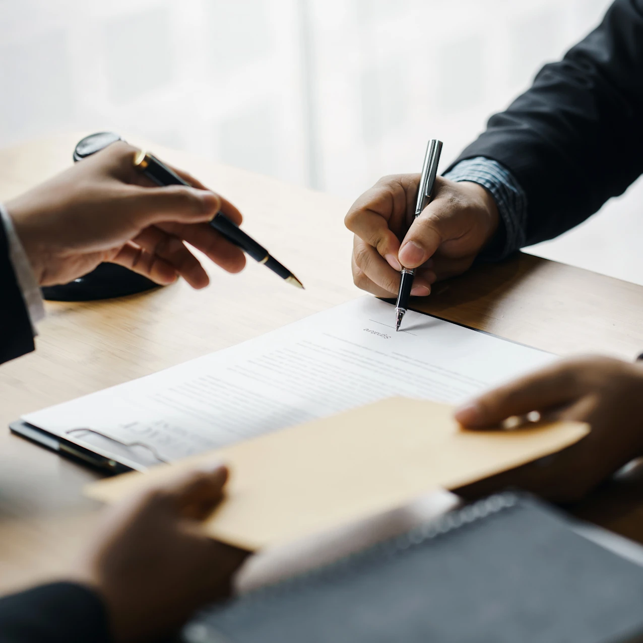 Two people reviewing and signing a document on a desk, one holding a pen and the other pointing, with an envelope and a notebook nearby.