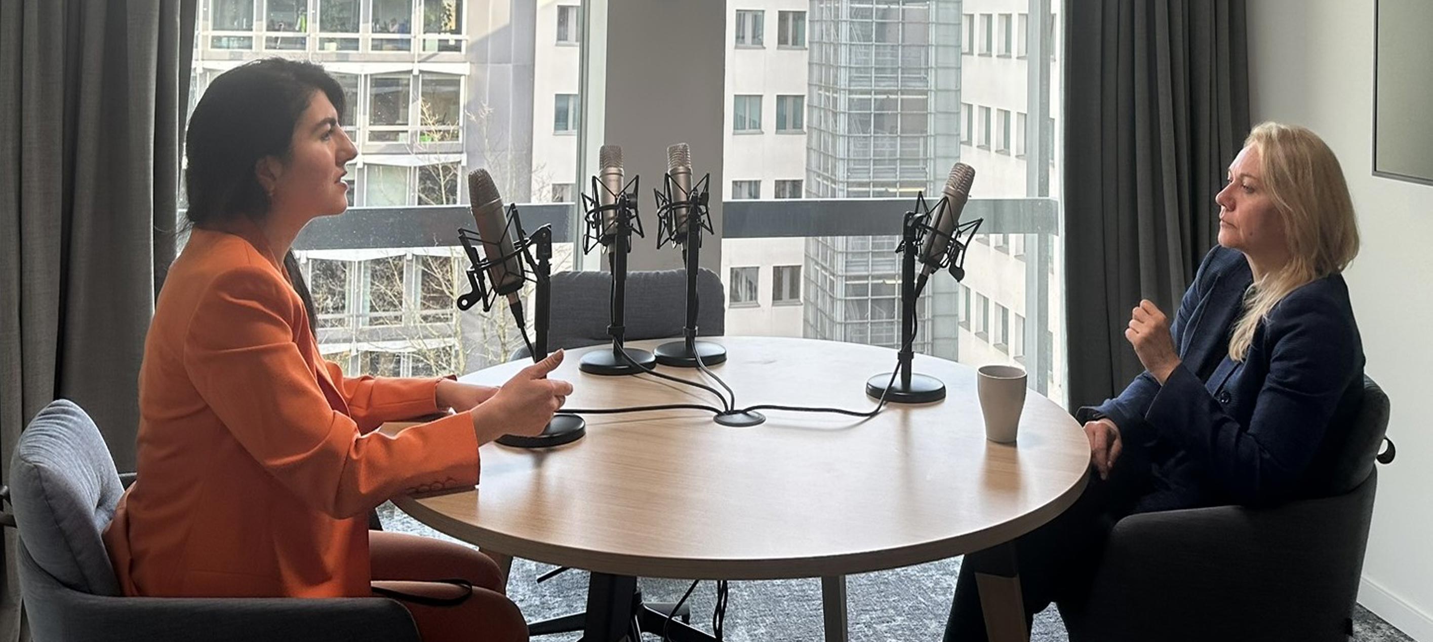 Two professionals sitting across from each other at a table with podcast mics in front of them