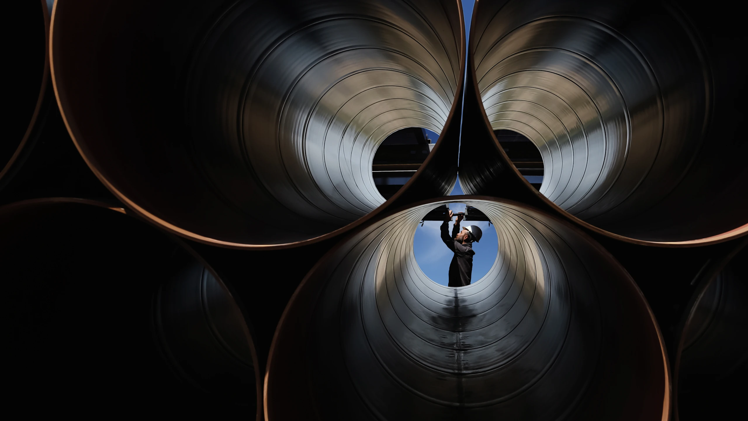 A worker wearing a helmet and safety glasses inspects large industrial metal pipes from inside one pipe, visible against a blue sky background.