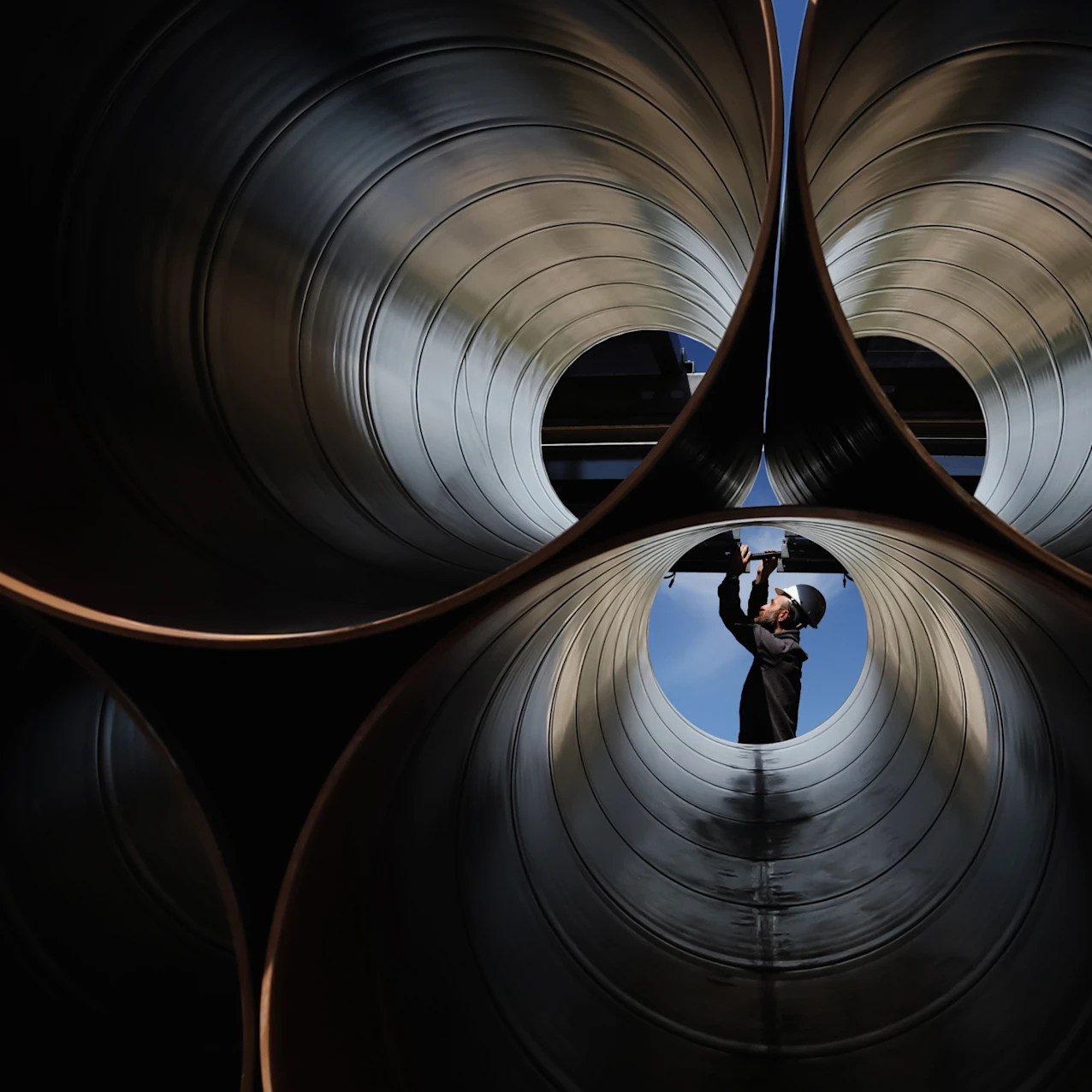 A worker wearing a helmet and safety glasses inspects large industrial metal pipes from inside one pipe, visible against a blue sky background.