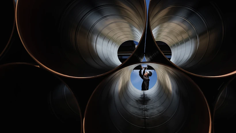 A worker wearing a helmet and safety glasses inspects large industrial metal pipes from inside one pipe, visible against a blue sky background.