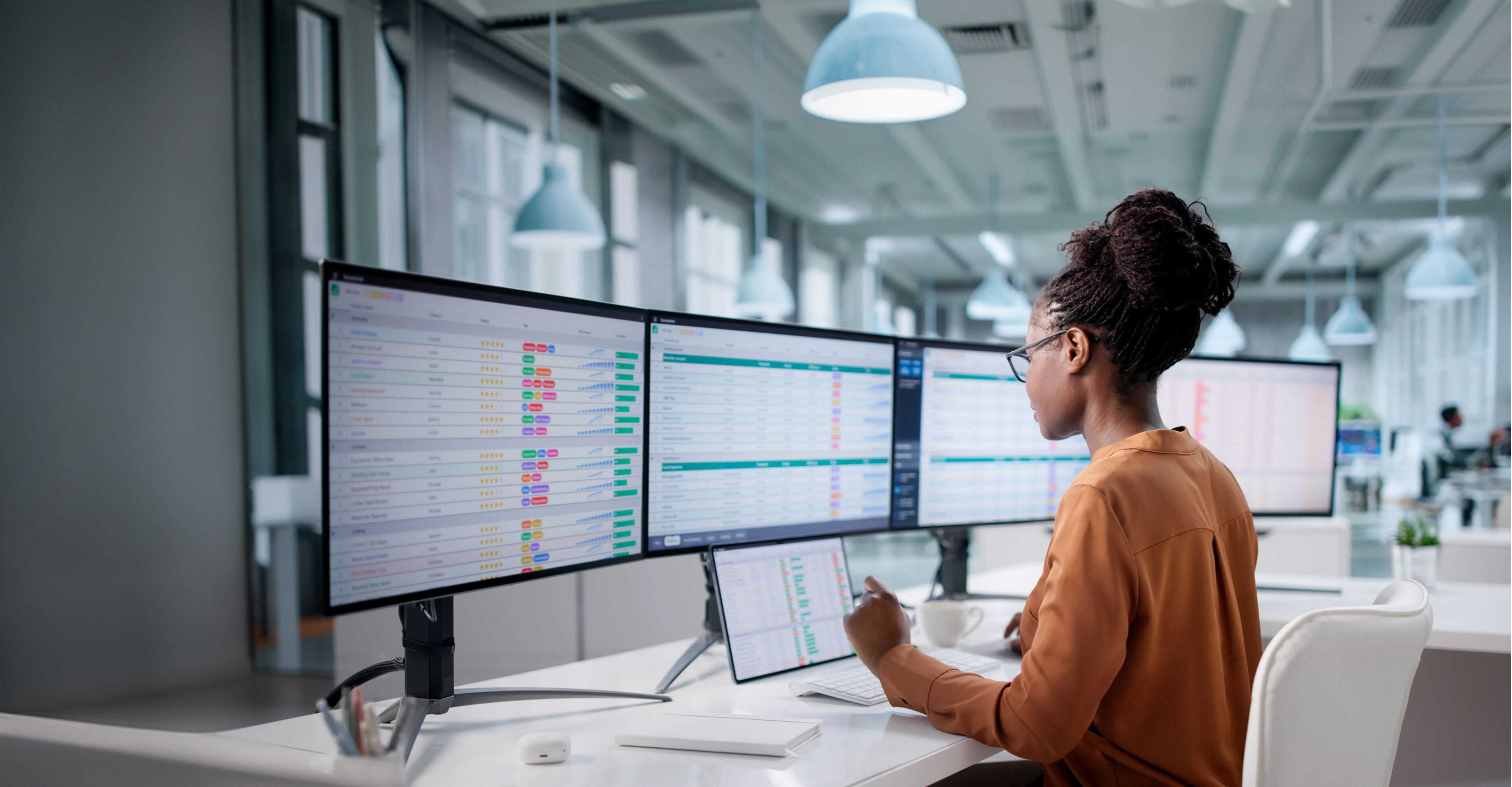A woman in glasses working at a desk with four large monitors displaying colorful charts and data in a modern office space with hanging lights.