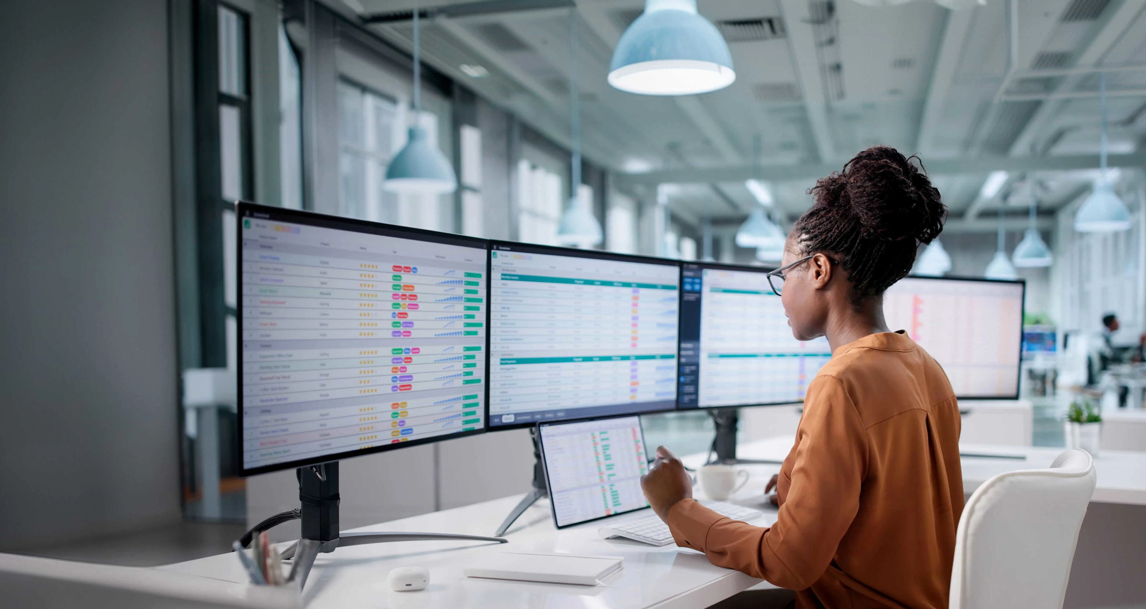A woman in glasses working at a desk with four large monitors displaying colorful charts and data in a modern office space with hanging lights.