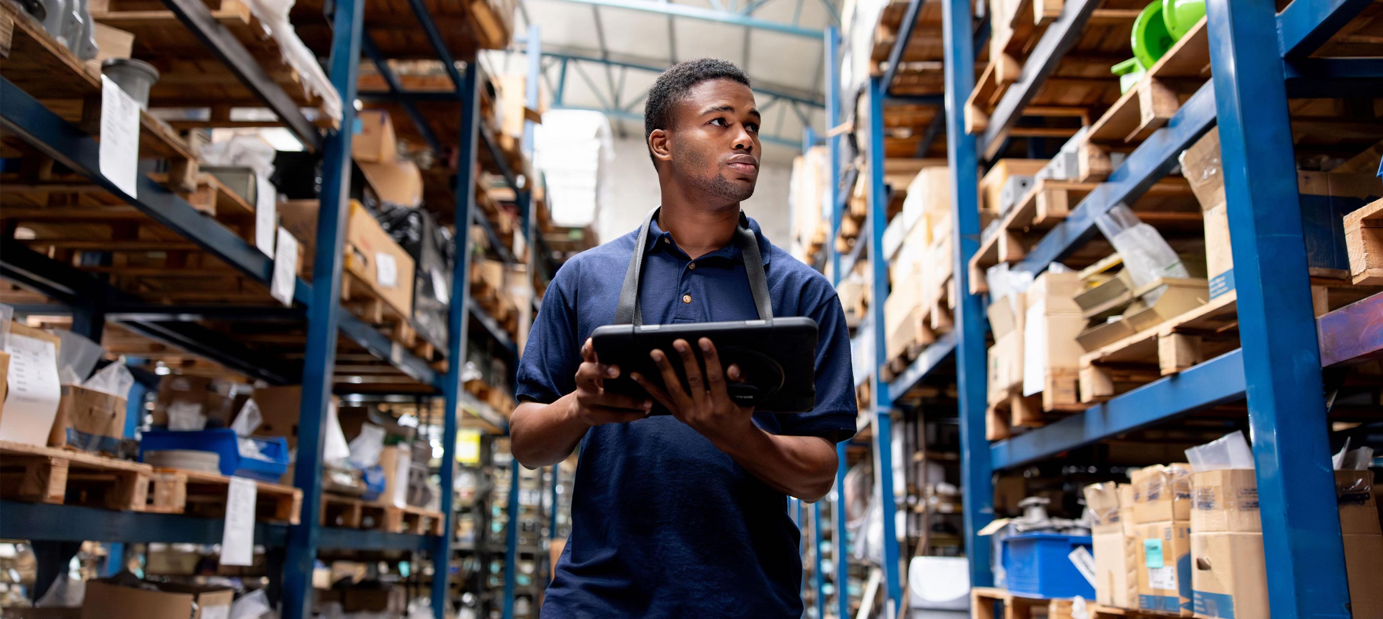 A man in a dark blue polo shirt holds a tablet and looks to the side while standing between tall warehouse shelves filled with boxes and packages.