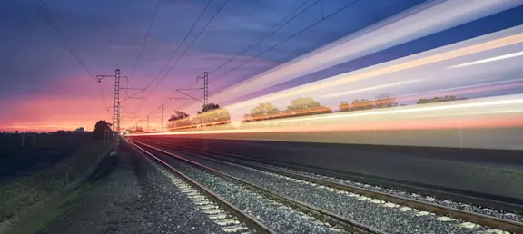 A high-speed train moves along railway tracks at sunset, leaving colourful light trails beneath power lines.