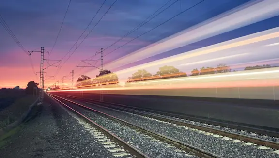 A high-speed train moves along railway tracks at sunset, leaving colourful light trails beneath power lines.