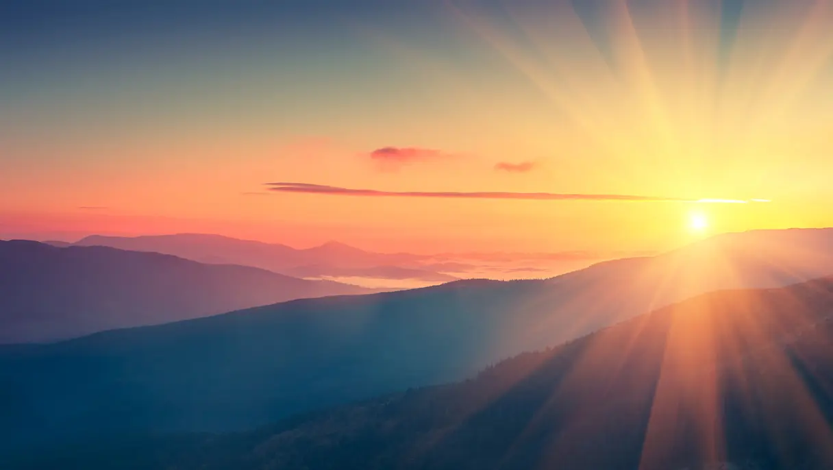 Sunrise over layered mountain silhouettes with mist in the valley and sun rays across an orange and blue sky.