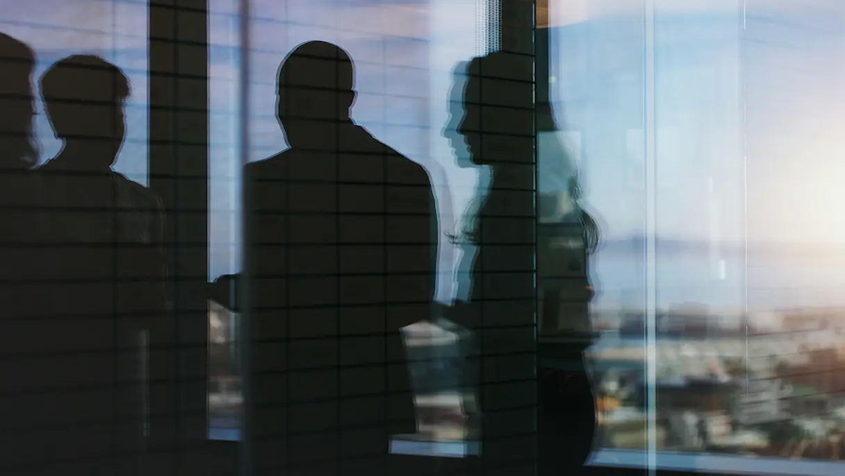 Silhouettes of four people behind a glass panel with a grid pattern, with a blurred city skyline visible beyond.
